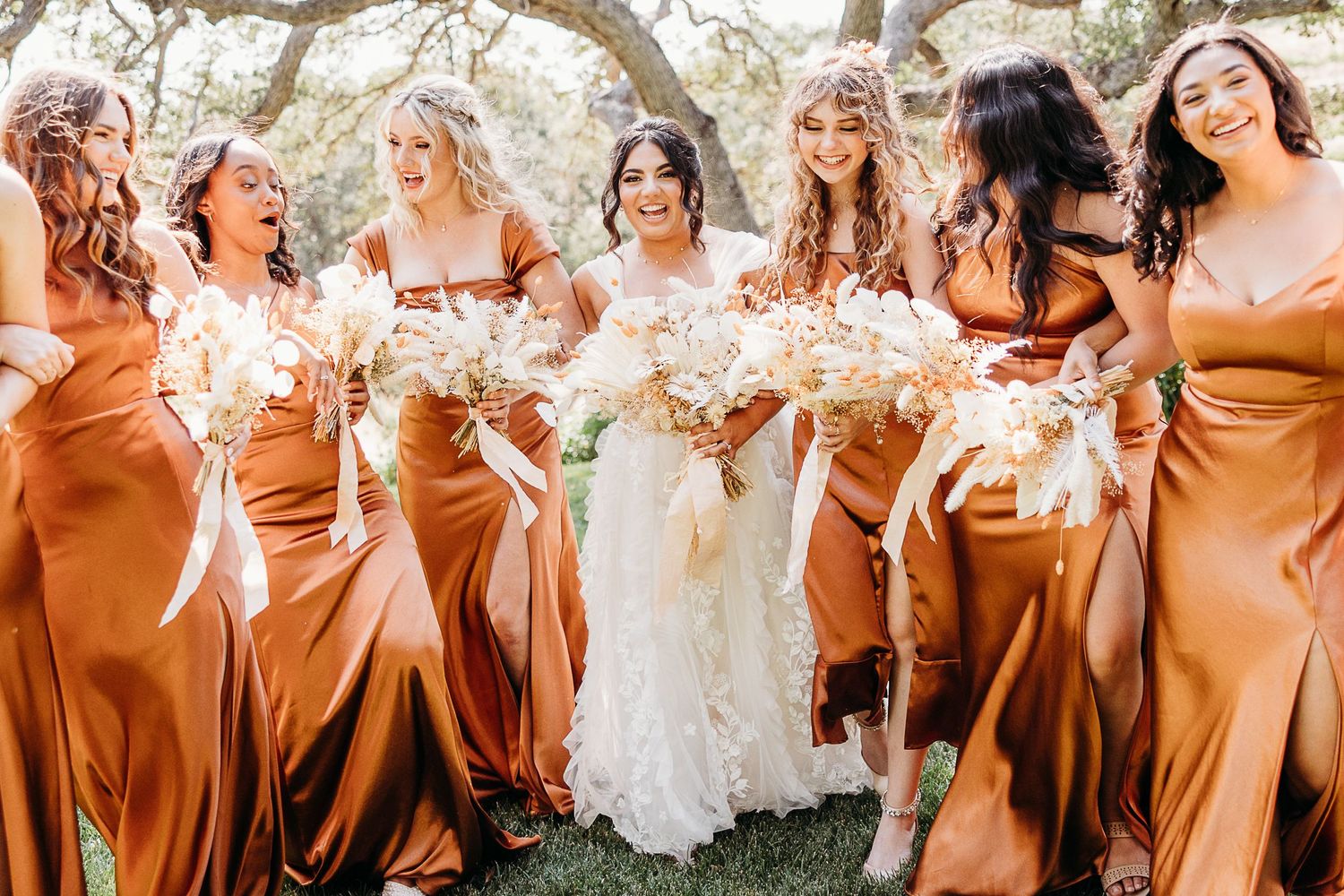 Candid shot of bridal party in rust silk dresses laughing together during outdoor wedding photos.