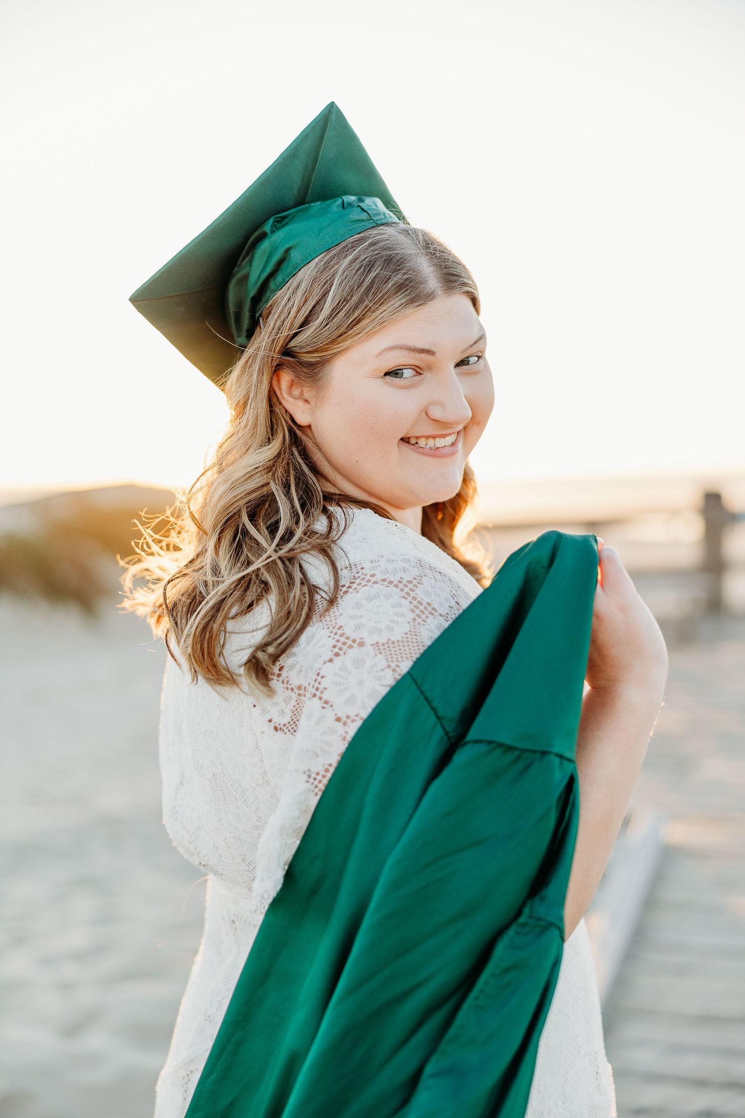 A graduate holds their green graduation gown while posing for a portrait at sunset.