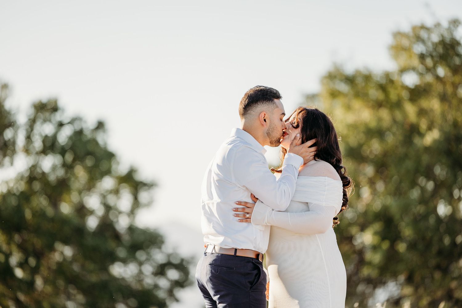 A couple shares a romantic kiss in a sunlit outdoor setting with trees in the background.