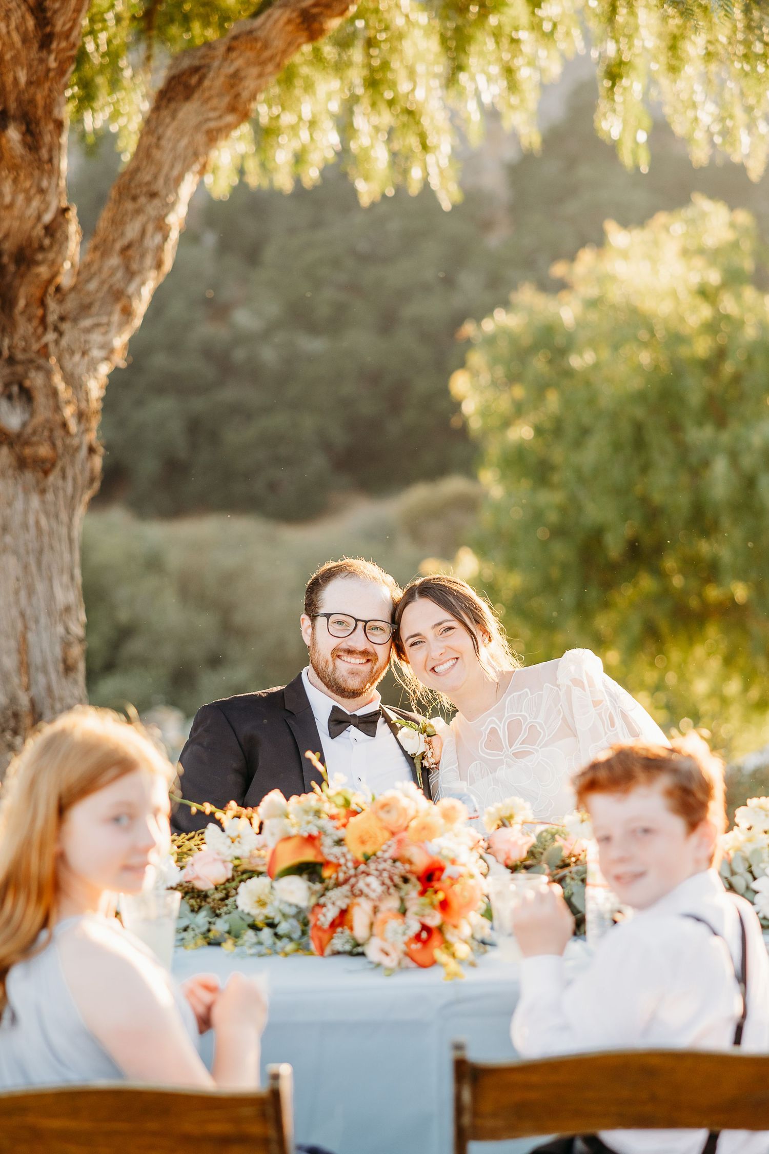 A couple enjoys their outdoor wedding dinner under a beautiful tree with orange floral centerpieces at sunset.