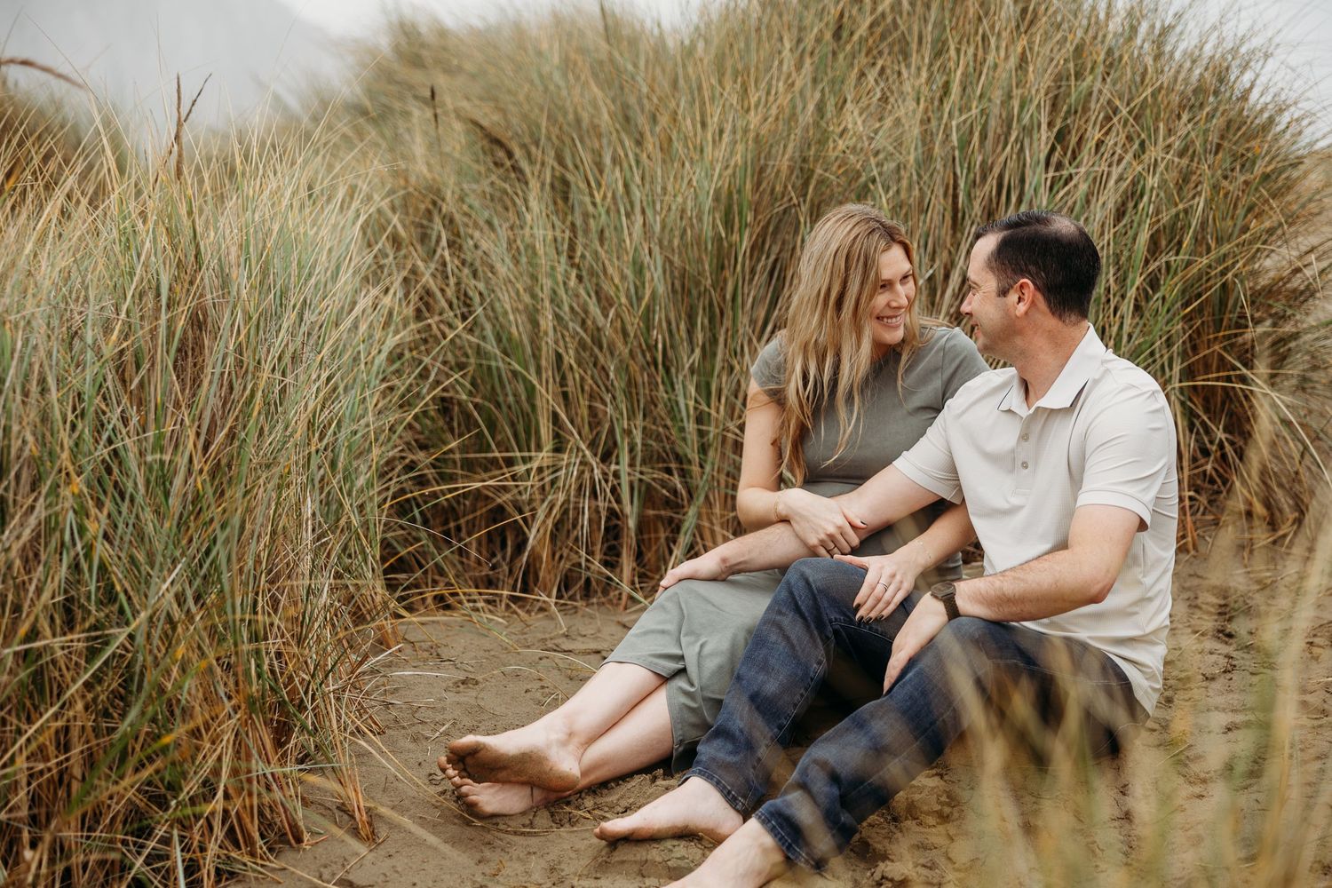 Casual outdoor maternity photo of couple relaxing together against coastal grass backdrop.
