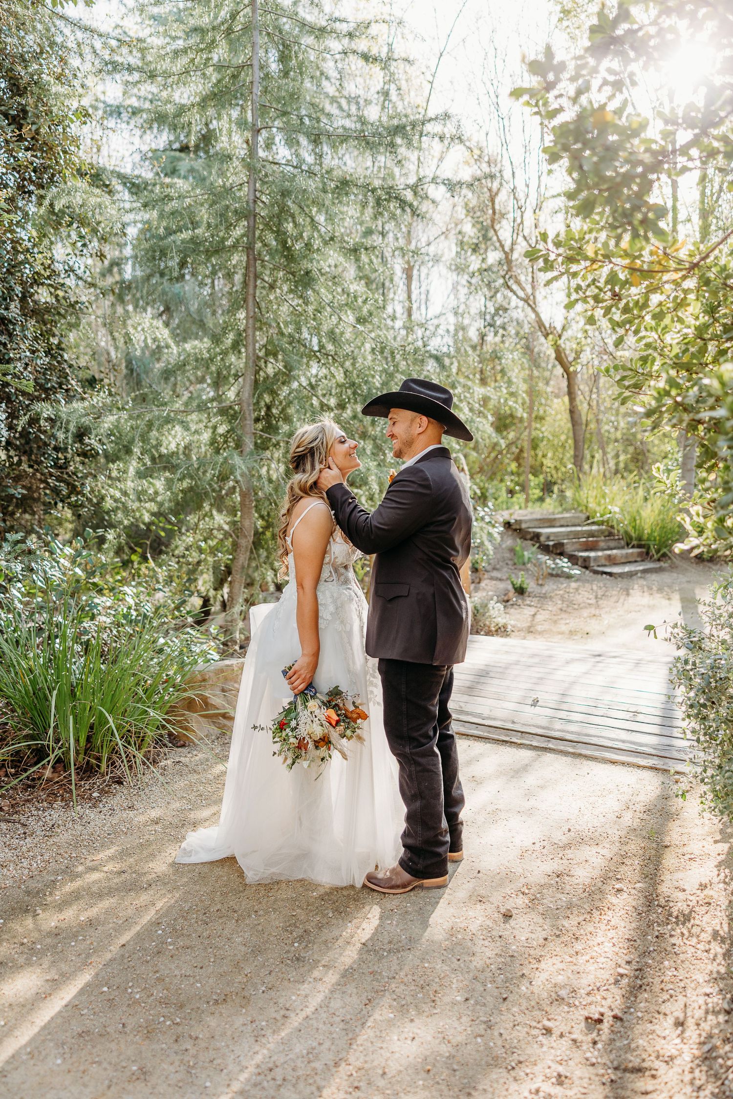 A couple shares a romantic moment on a sunlit woodland path while dressed in wedding attire.