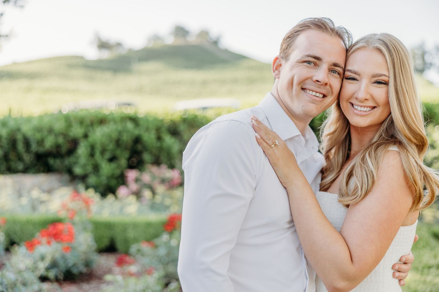 A couple poses together in an outdoor engagement photo session with rolling hills and wildflowers in the background.