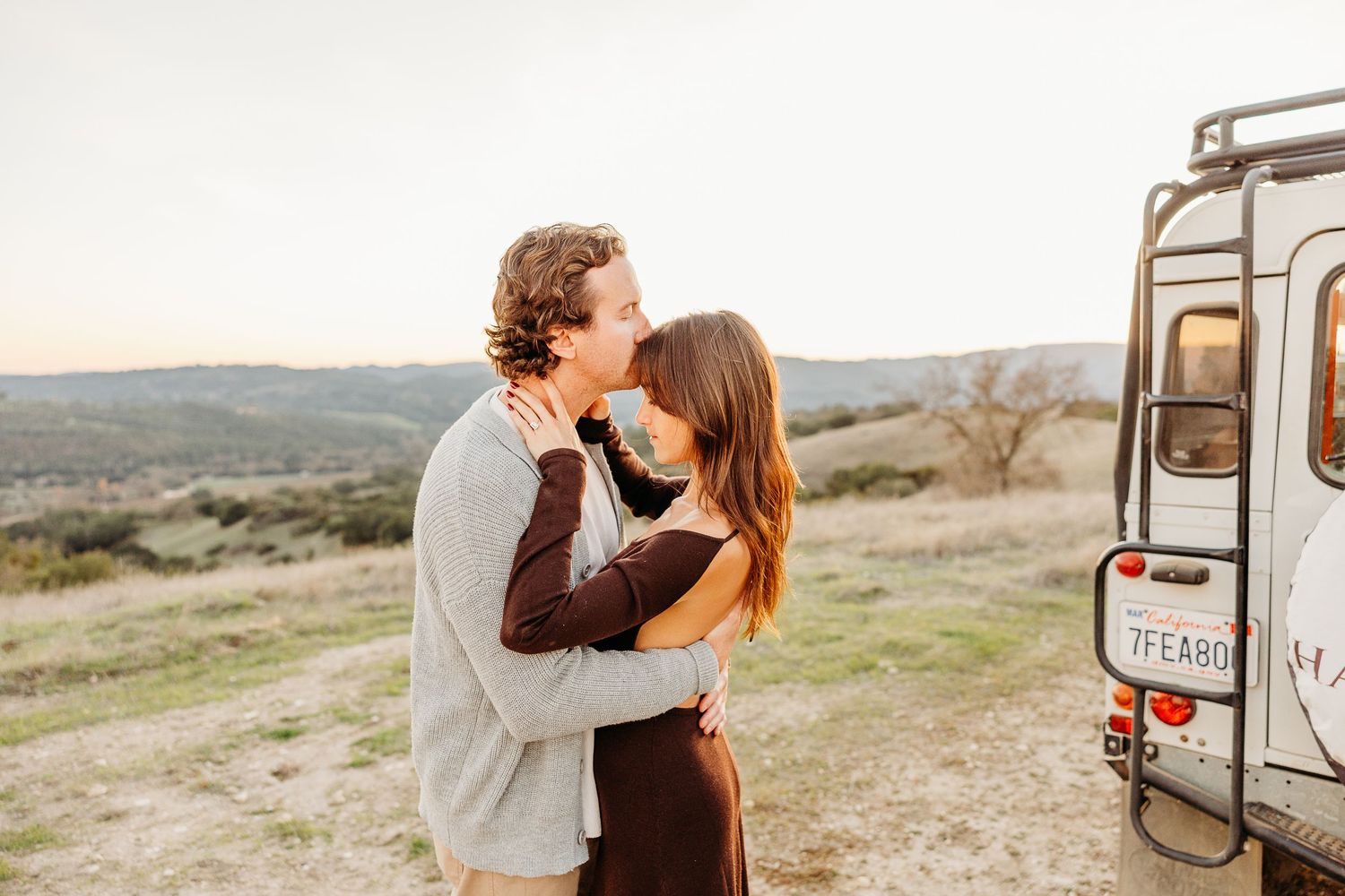Two people share romantic moments near a parked Land Rover with mountains in the distance.