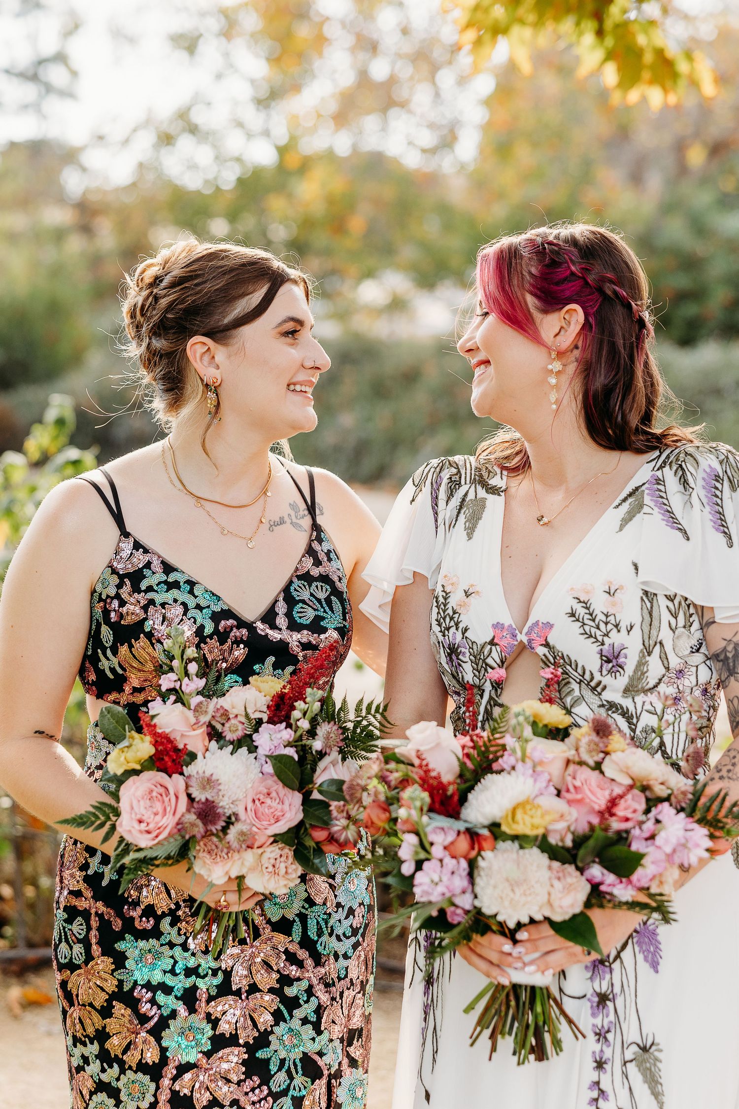 Wedding couple holding bouquets of pink and red flowers while laughing together outdoors.