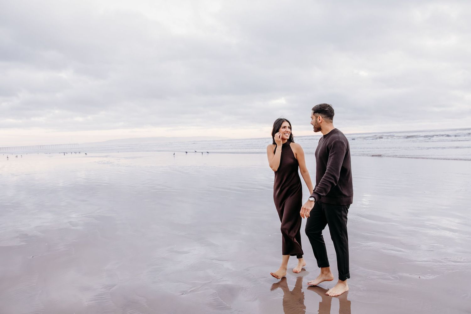 Couple stands barefoot on reflective beach during overcast day.