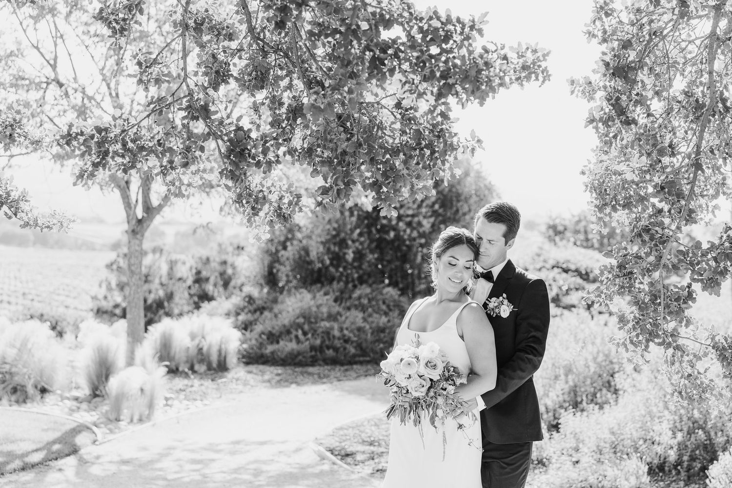 A black and white wedding photo of a couple embracing outdoors in a garden setting with trees and natural scenery.