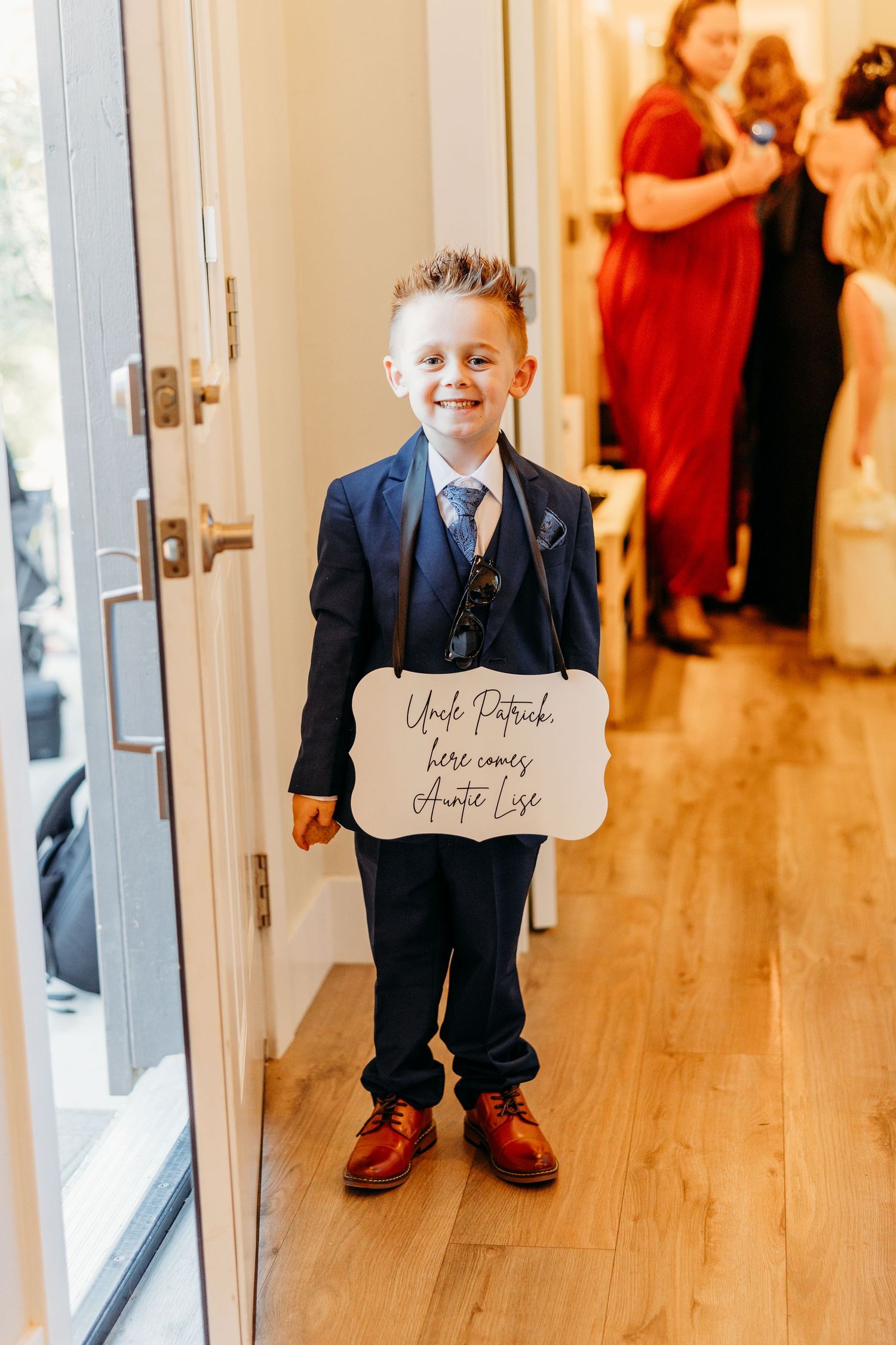 A young ring bearer in a navy suit holds a sign while standing in a bright hallway.