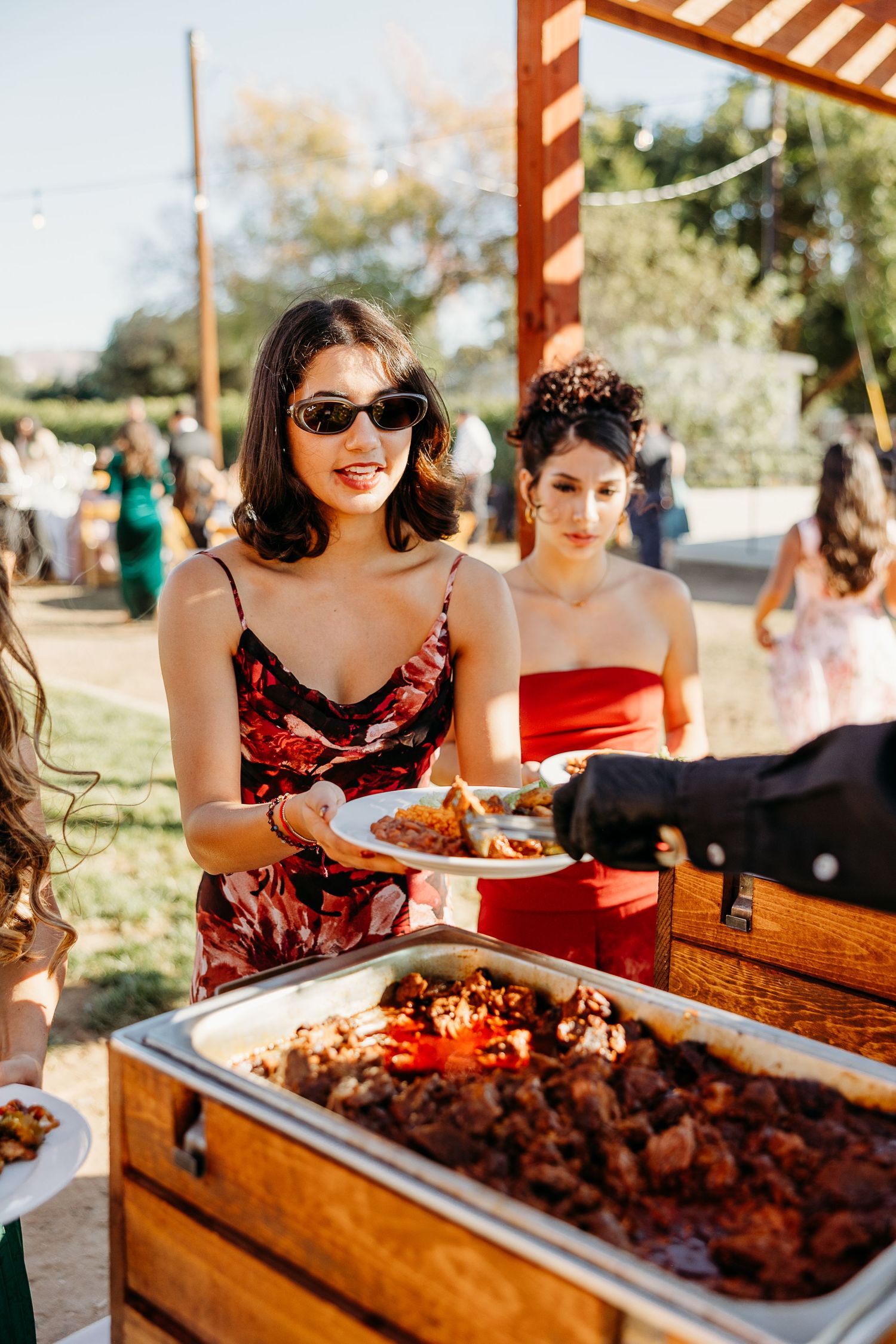 Wedding guests in vintage-style dresses serving food at an outdoor buffet table.
