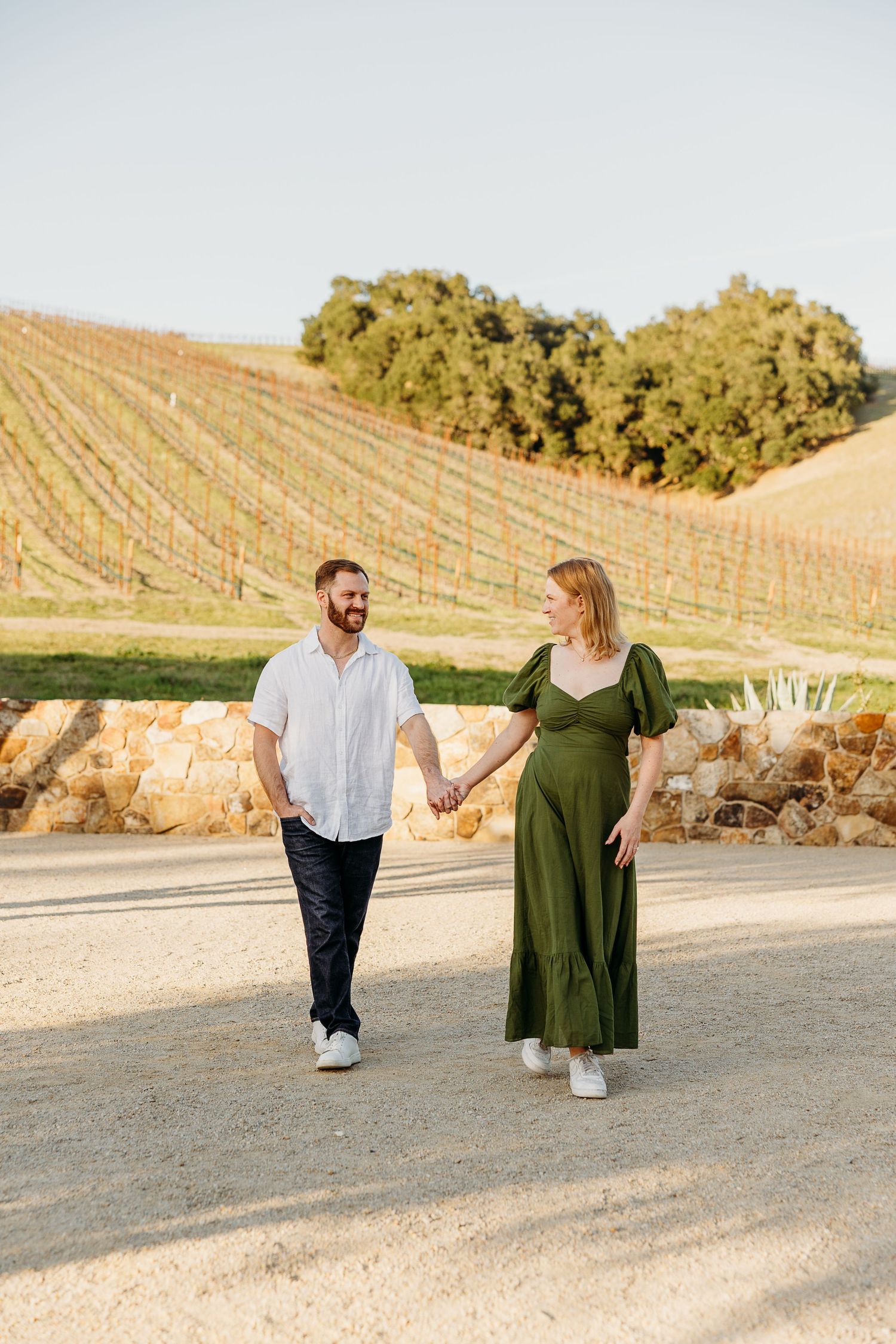 A couple takes a romantic stroll through a vineyard during golden hour while holding hands.
