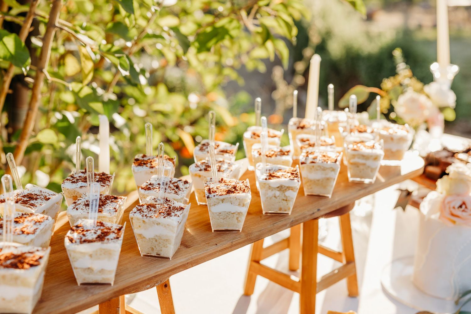 Rows of elegant takeout boxes filled with desserts line a rustic wooden table at an outdoor event.