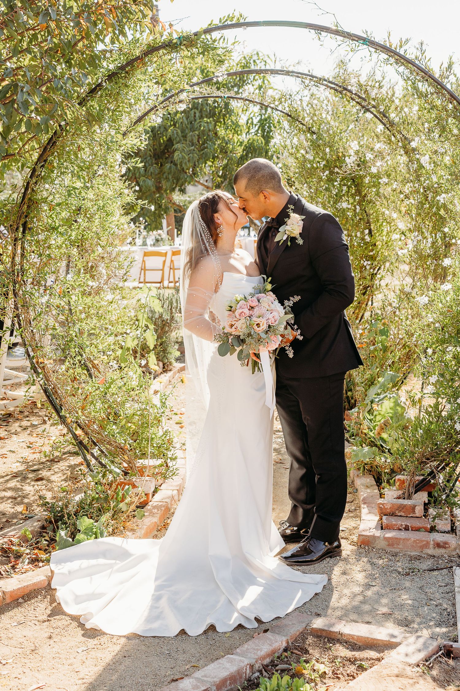 A romantic wedding portrait of a couple kissing under an arched garden trellis with greenery.