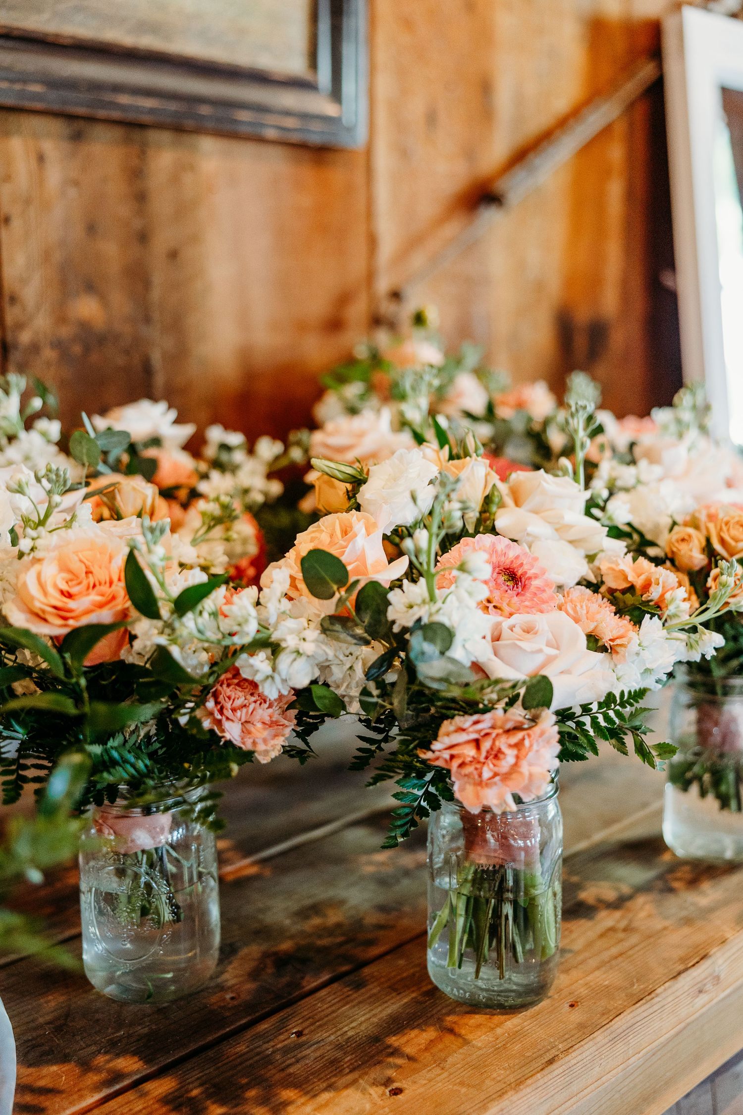 Mason jars filled with peach and pink floral arrangements line a rustic wooden table for wedding decor.