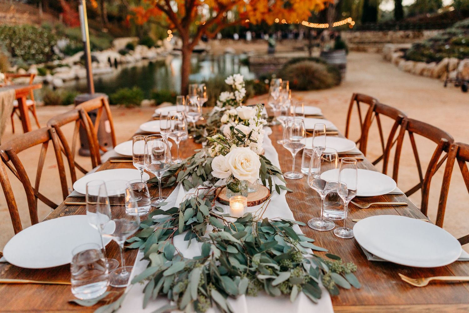 Elegant outdoor wedding reception table settings with rustic wooden chairs and eucalyptus centerpieces at sunset.