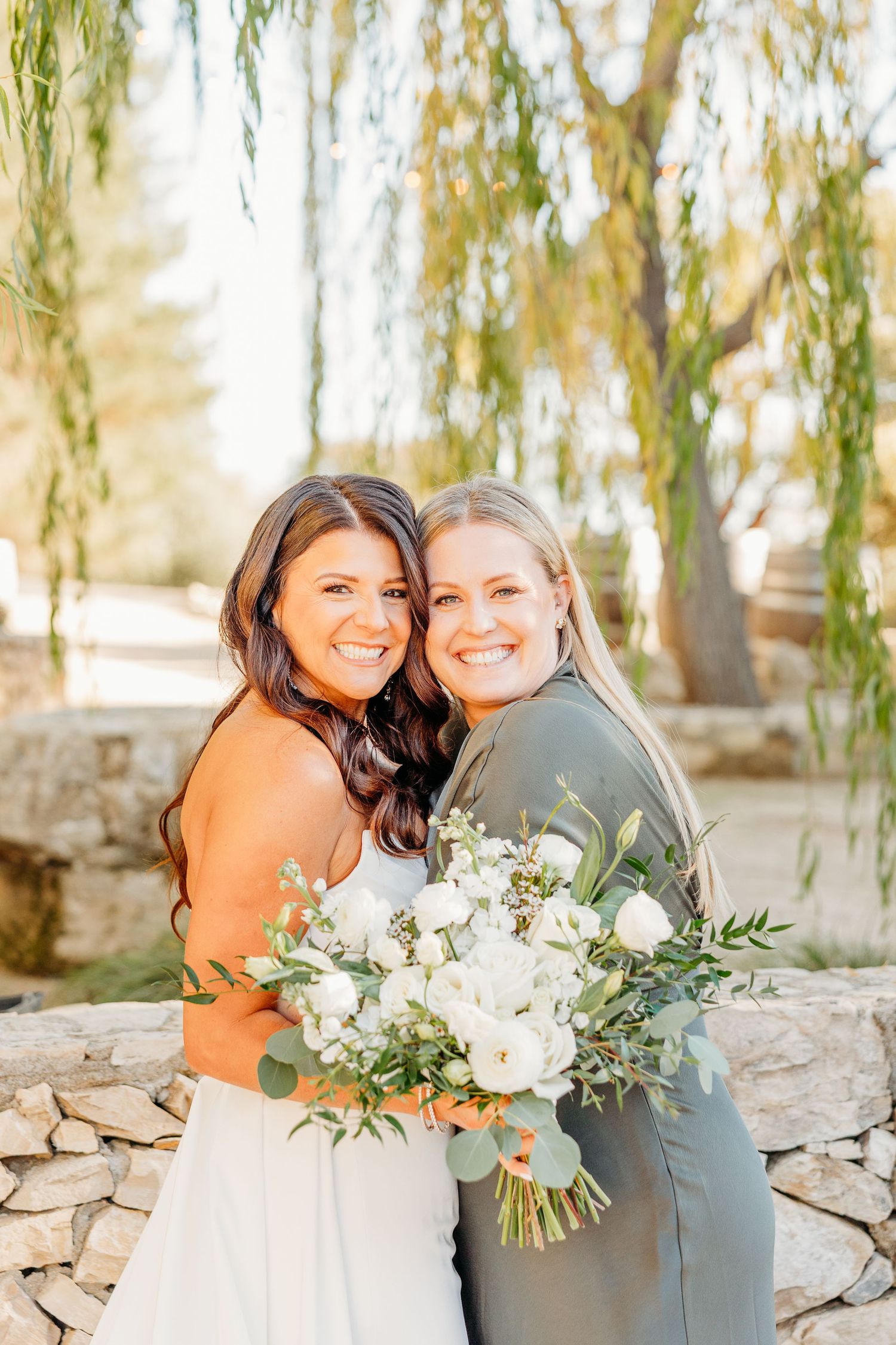 Wedding party members hold white floral bouquets against stone wall backdrop.