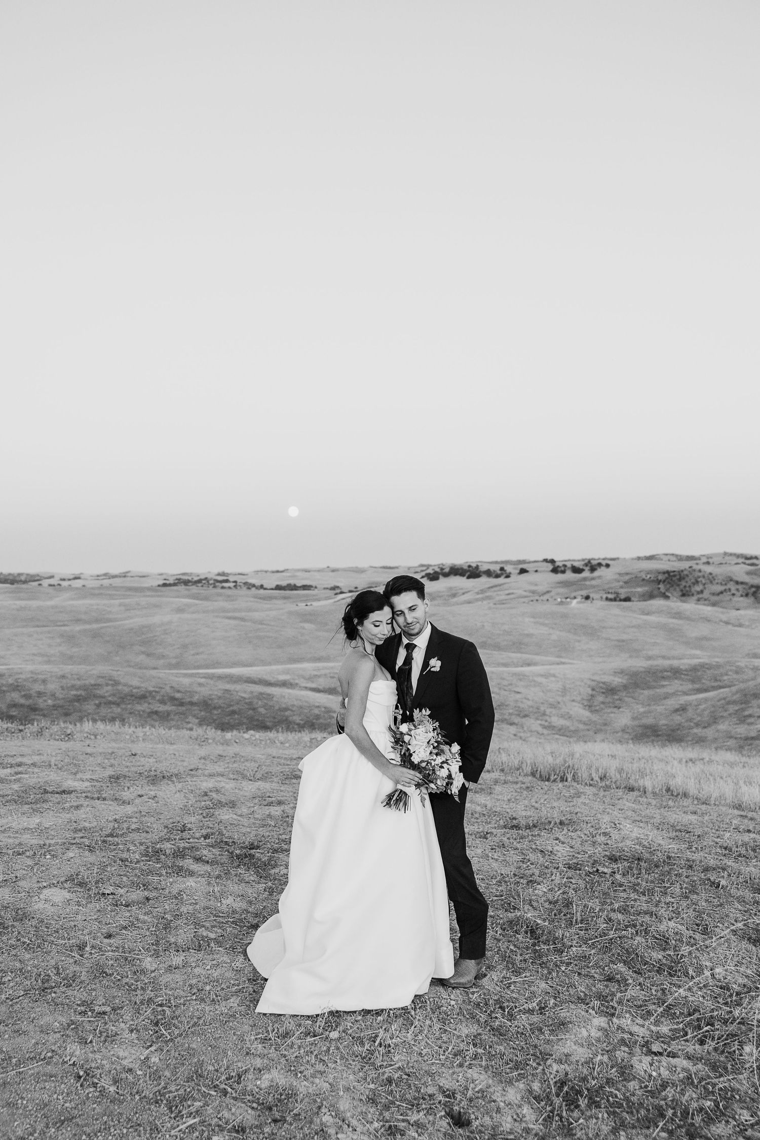 Black and white portrait of newlyweds embracing in an open field with rolling hills in the background.