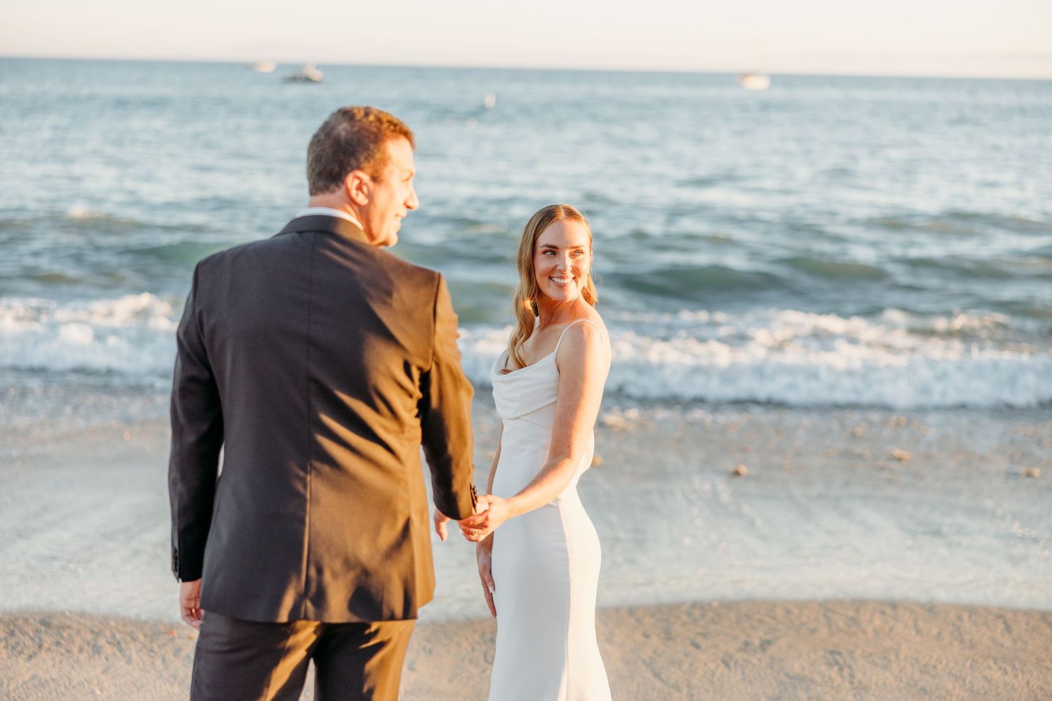 A couple in wedding attire standing together on a beach at sunset with ocean waves in the background.