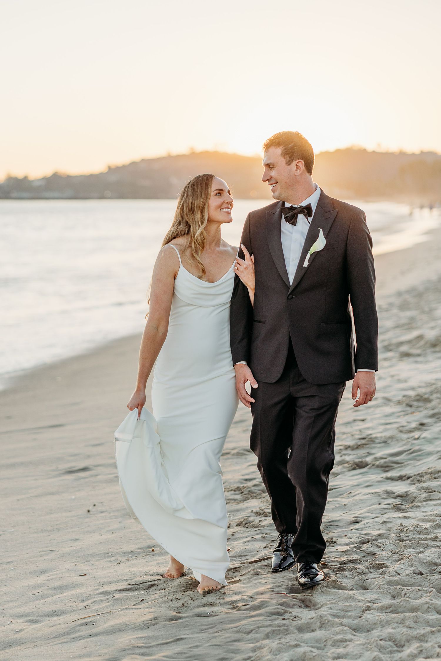 Couple walks hand in hand along a sunset beach in formal wedding attire.