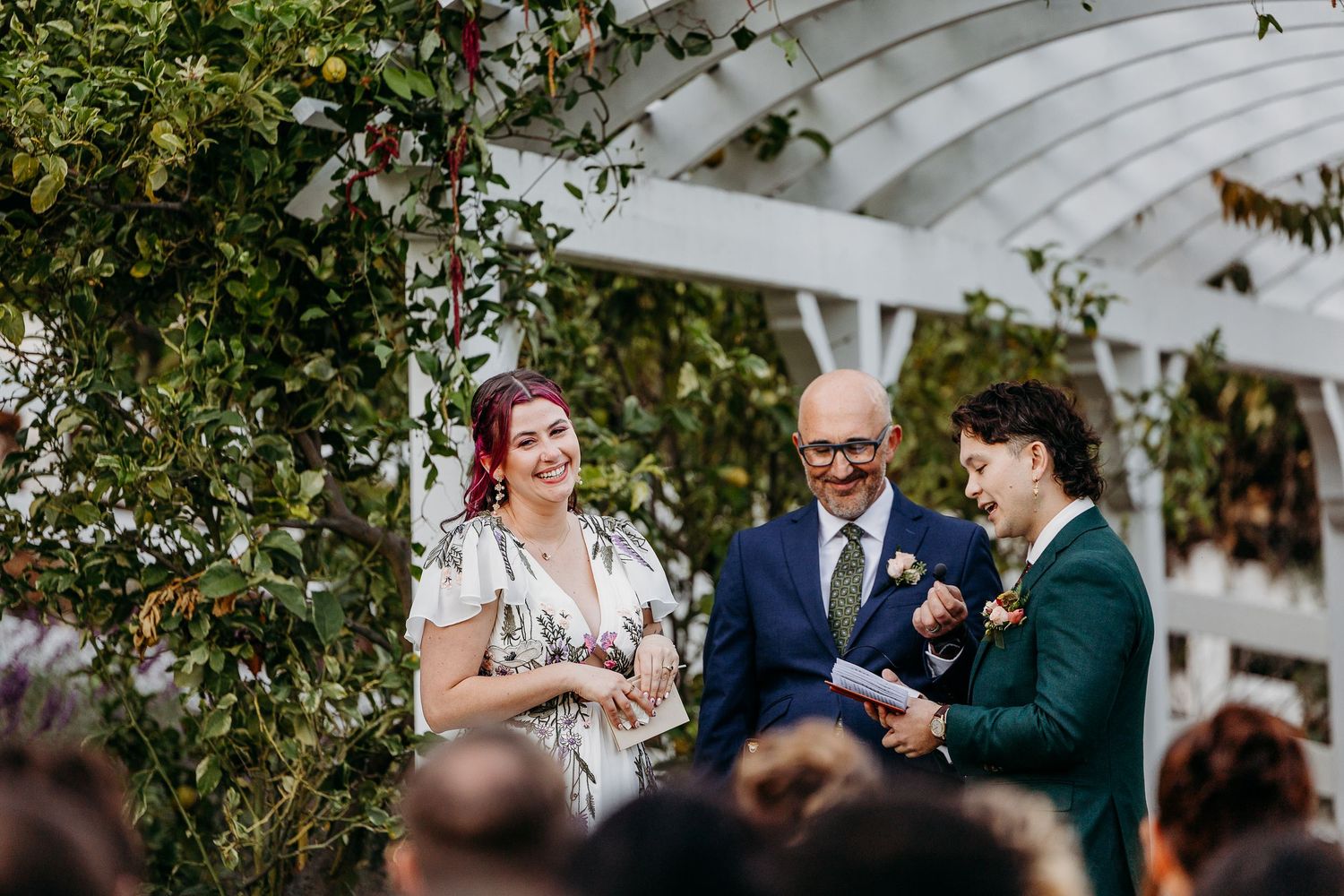 Wedding ceremony taking place under a white garden arbor surrounded by autumn trees.