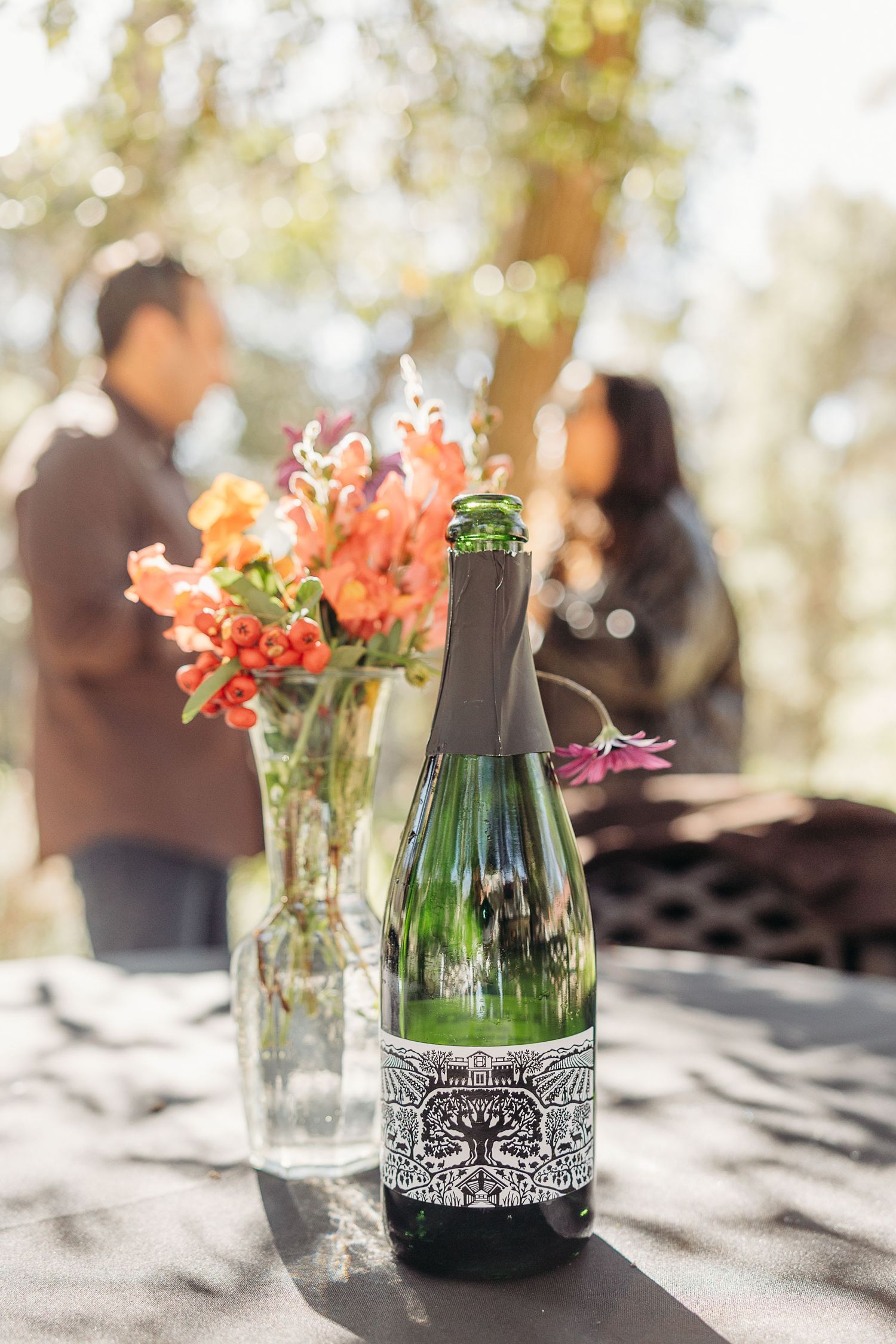 Wine bottle and floral arrangement on table with blurred figures in background.