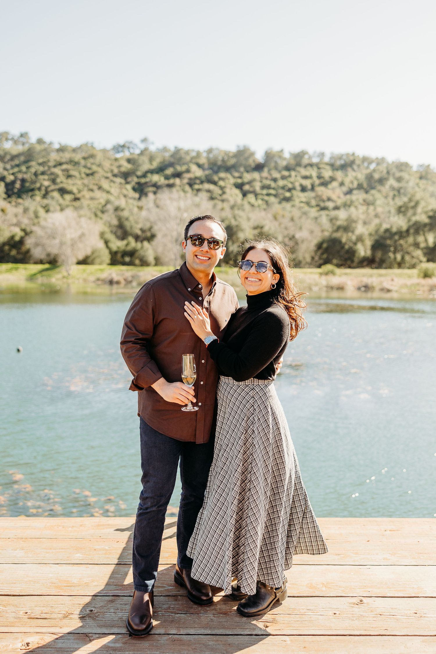 A couple shares a joyful moment together on a wooden dock overlooking a serene lake on a sunny afternoon.
