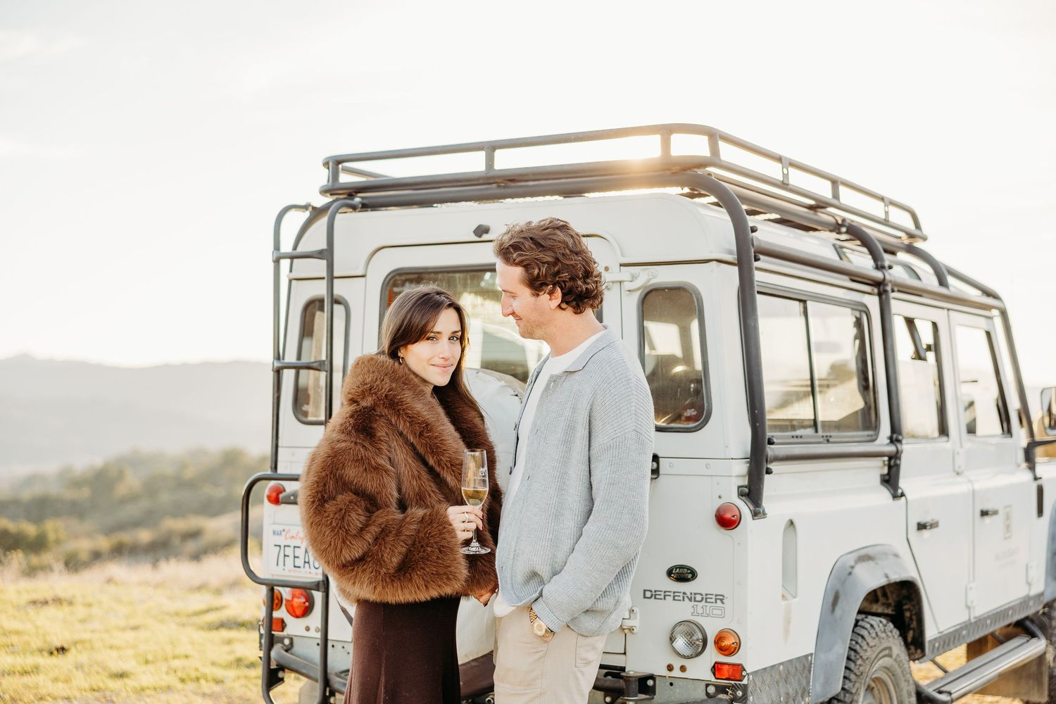 A romantic scene unfolds around a classic Land Rover parked in a scenic mountainous landscape.