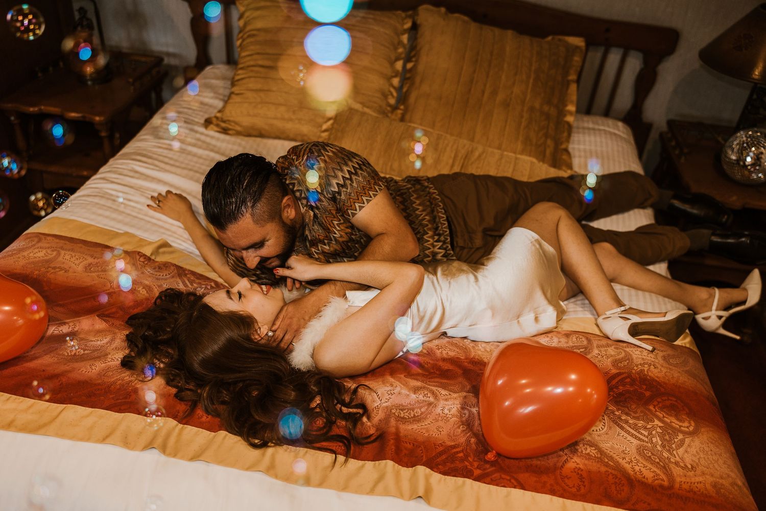 A romantic couple lies on a bed decorated with orange balloons and warm lighting creating an intimate atmosphere.
