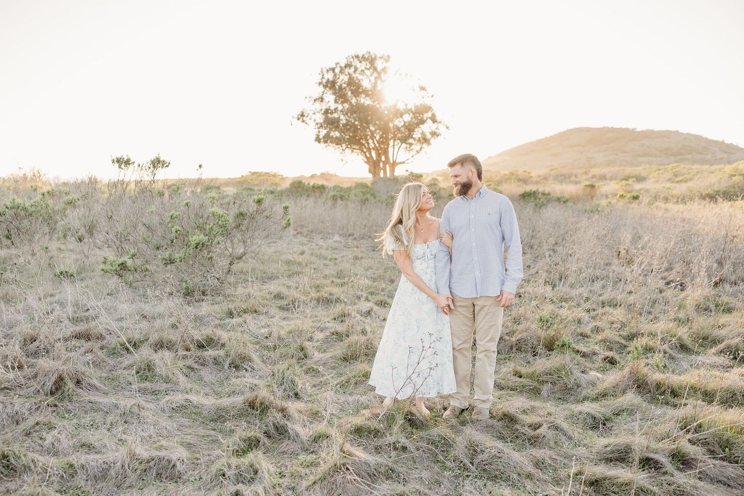 A romantic countryside engagement photo session with a couple walking through a golden sunlit field at sunset.