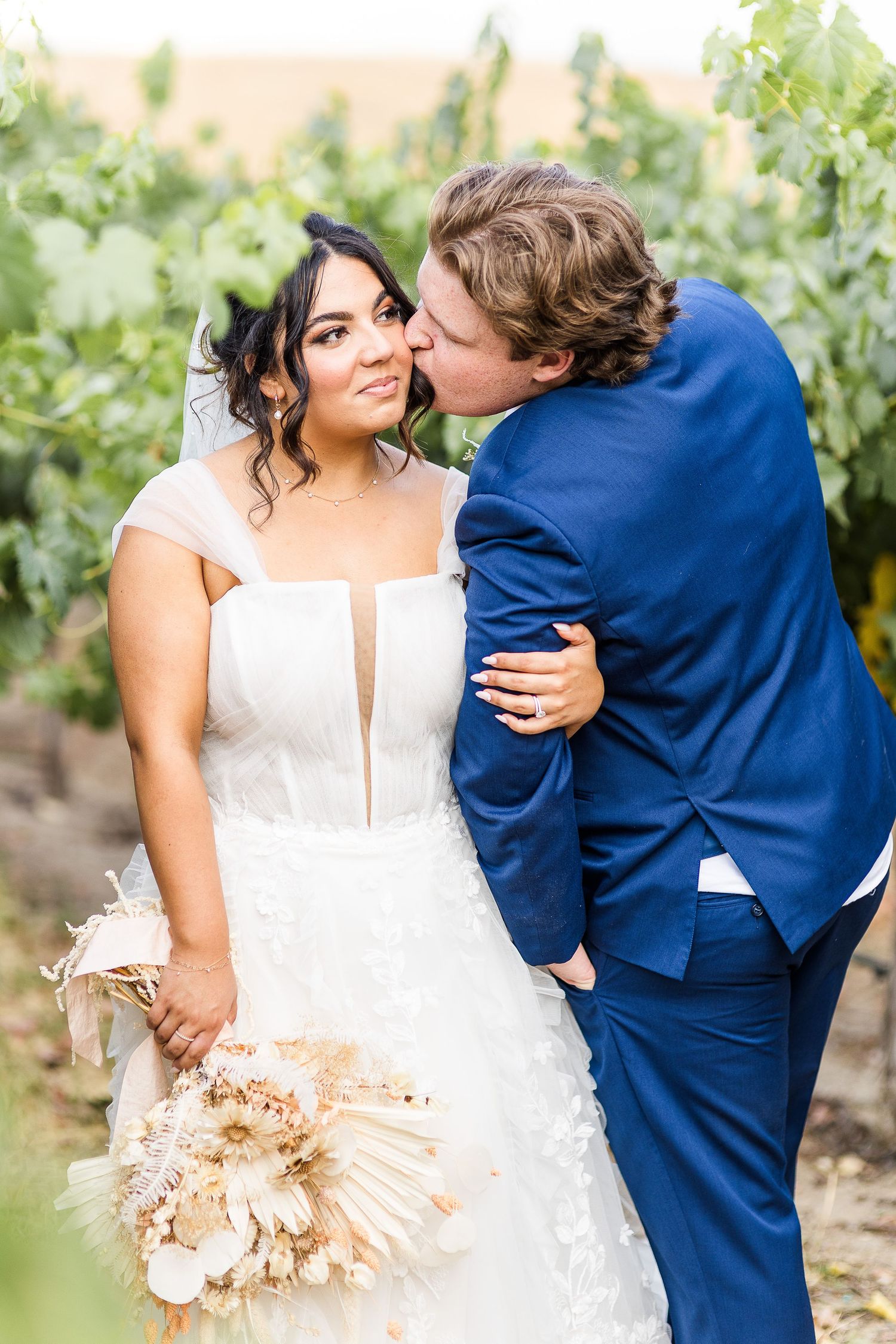 Tender moment between newlyweds in vineyard with bride holding neutral toned bouquet.