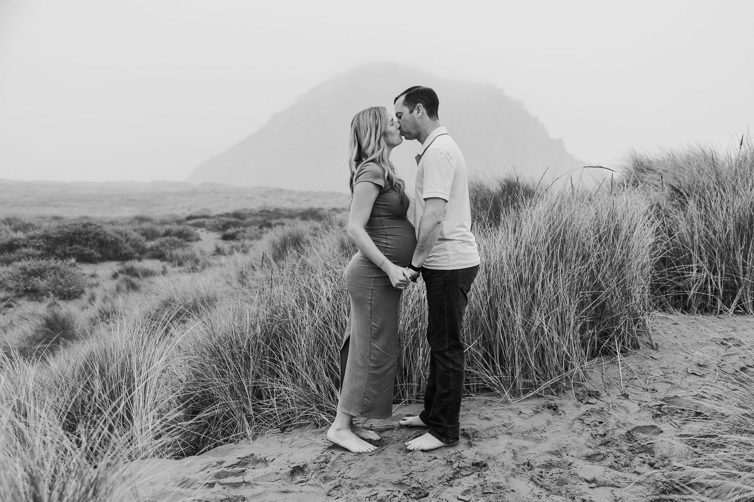 Black and white scenic maternity photo with couple embracing on sandy dunes.