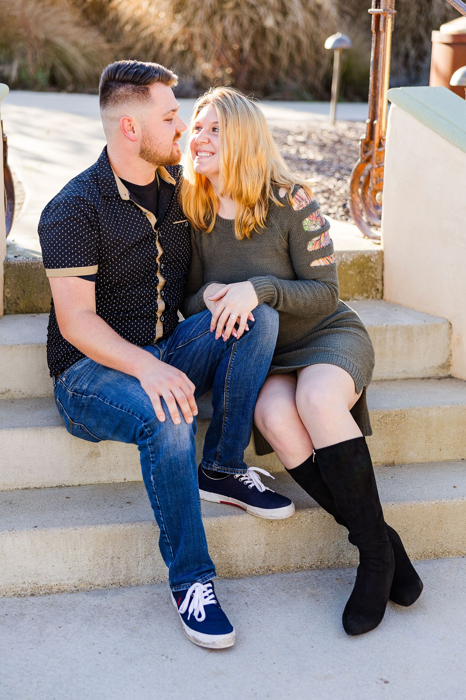 Couple sits close together on concrete steps with rustic building backdrop.