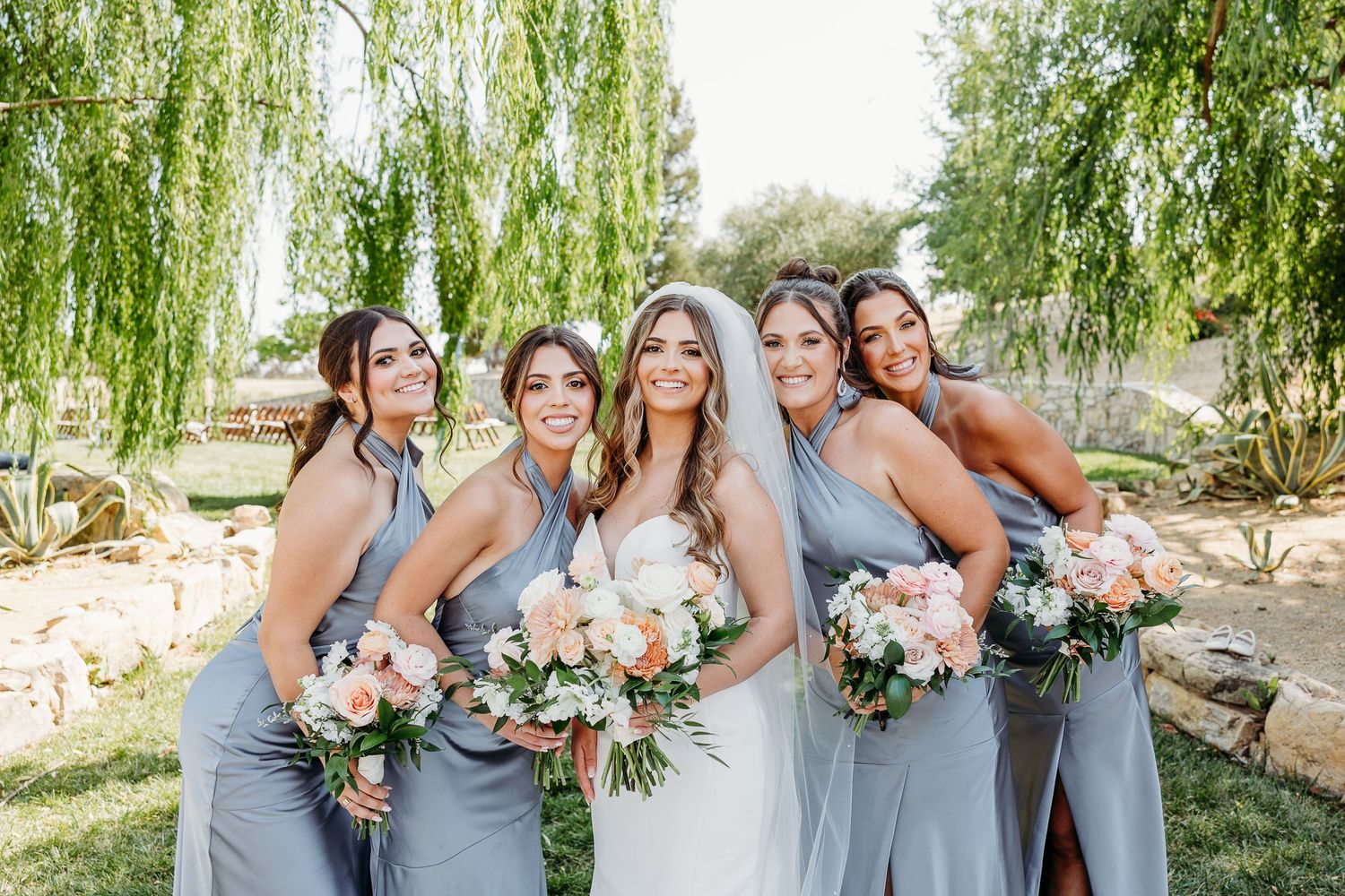 A bridal party in flowing grey dresses holds beautiful pastel bouquets under a weeping willow tree on a sunny day.