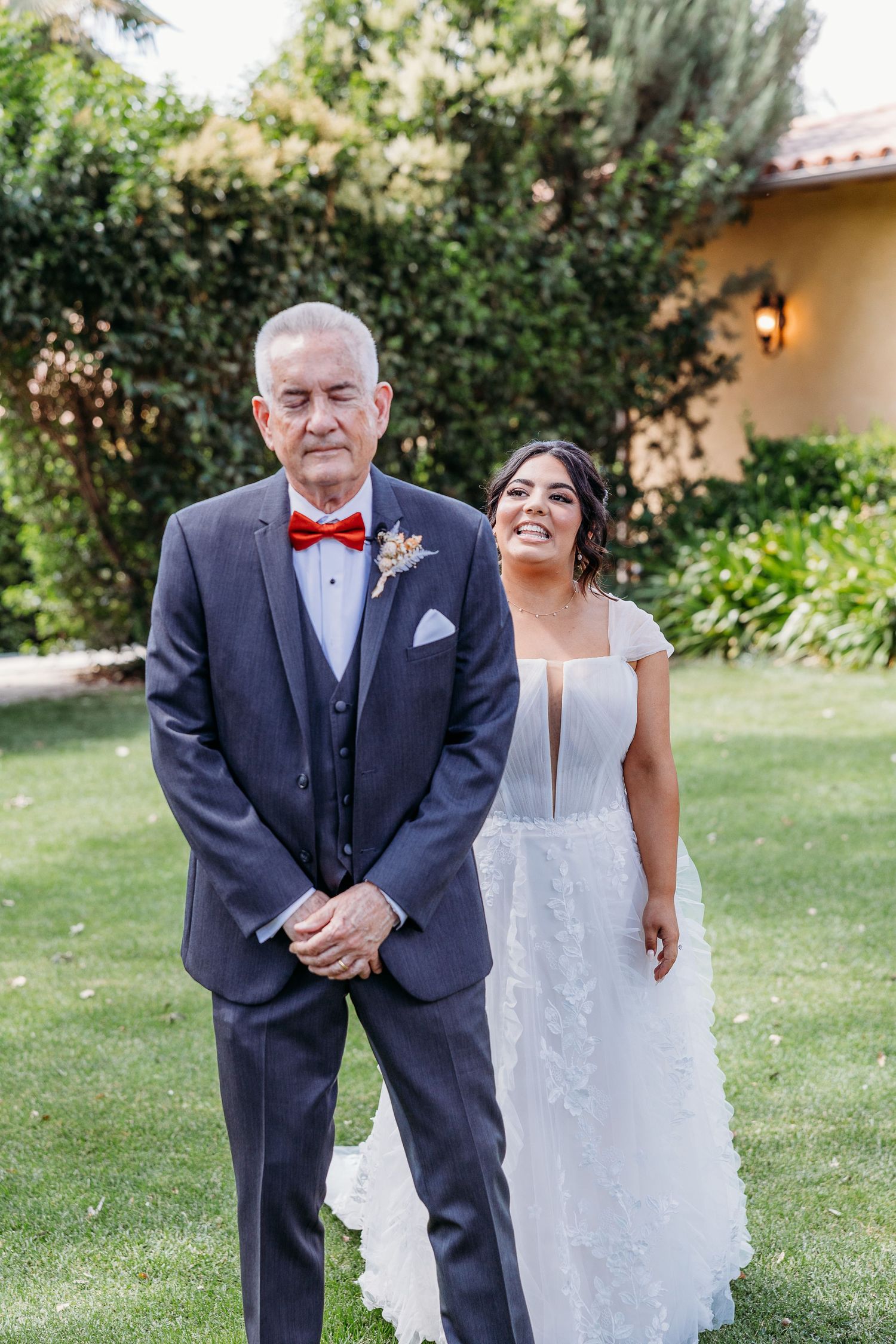 A couple poses in formal wedding attire outdoors, with the person in navy suit wearing a red bowtie.