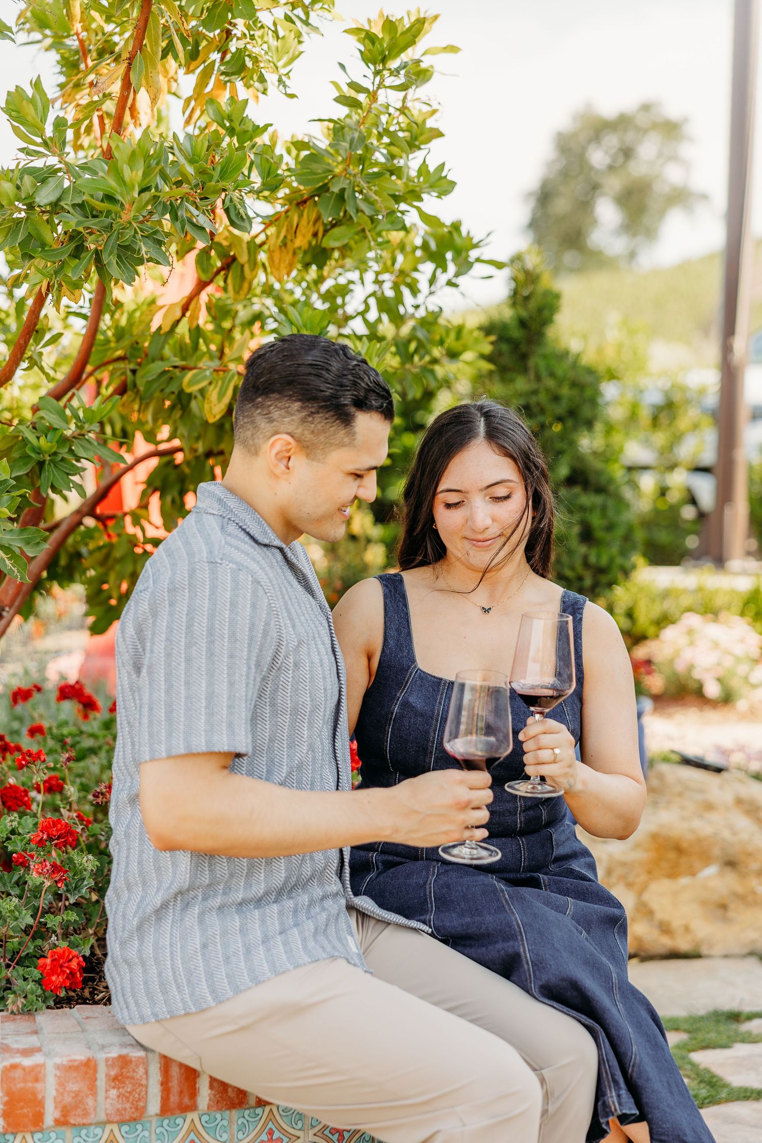 A couple enjoying wine together in a garden with red flowers and lush greenery.