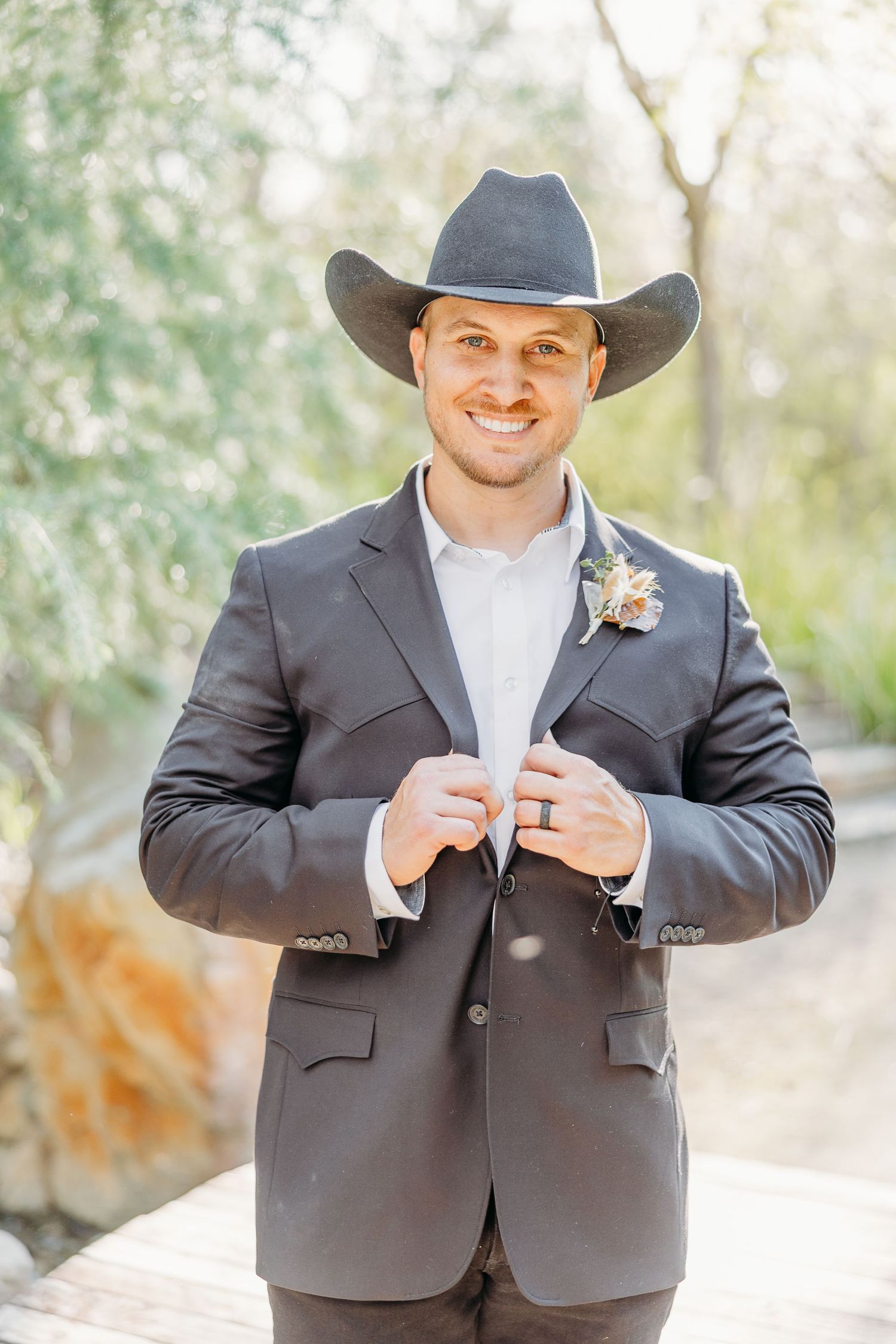 Groom in dark suit and cowboy hat smiles while adjusting jacket in outdoor setting.