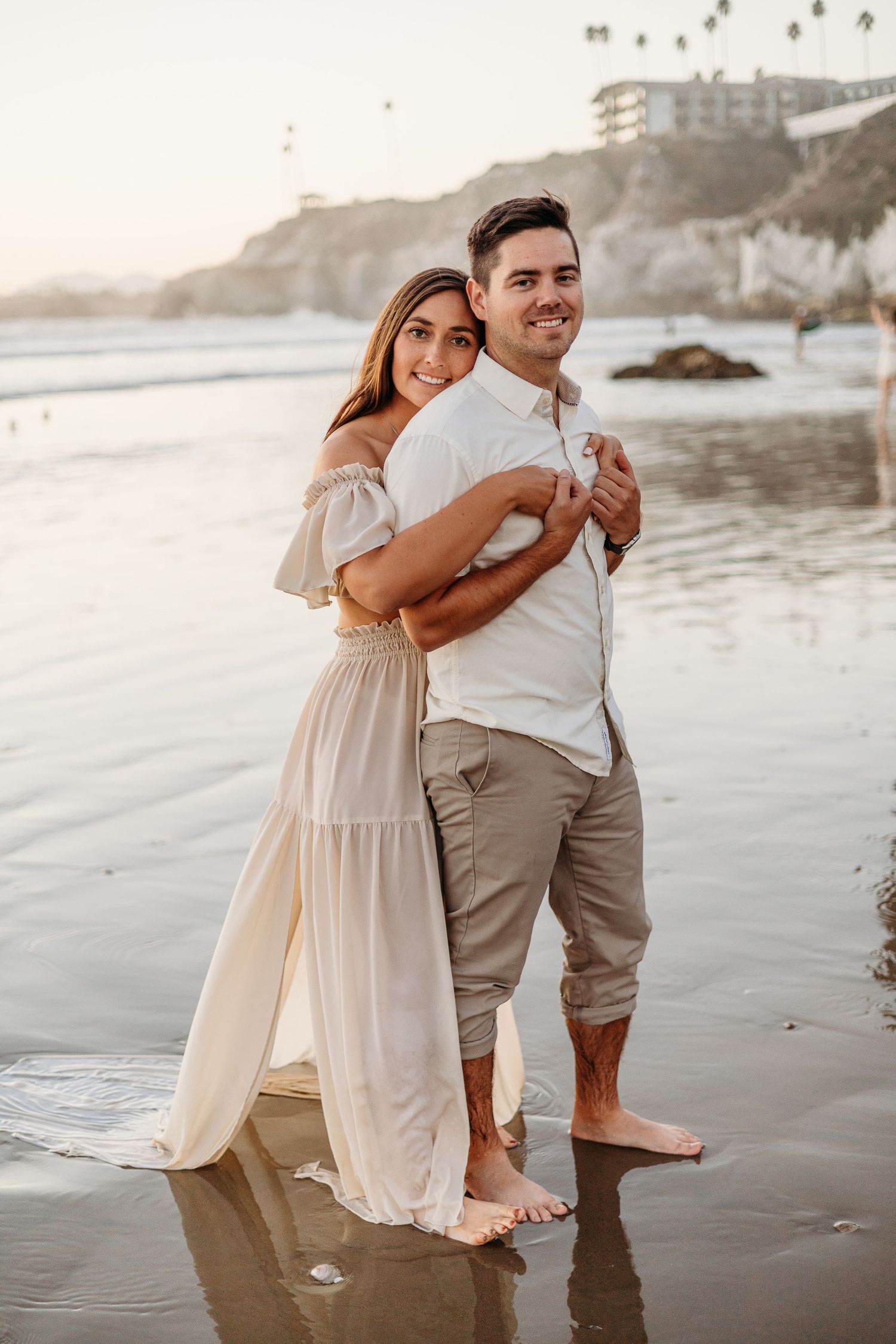 A couple embracing on a sandy beach with coastal cliffs and ocean waves during golden hour.