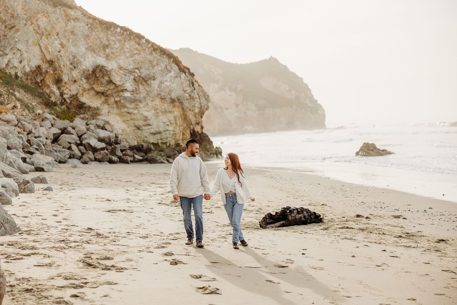Couple walking hand in hand on foggy beach with dramatic cliffs and rocky coastline in background.