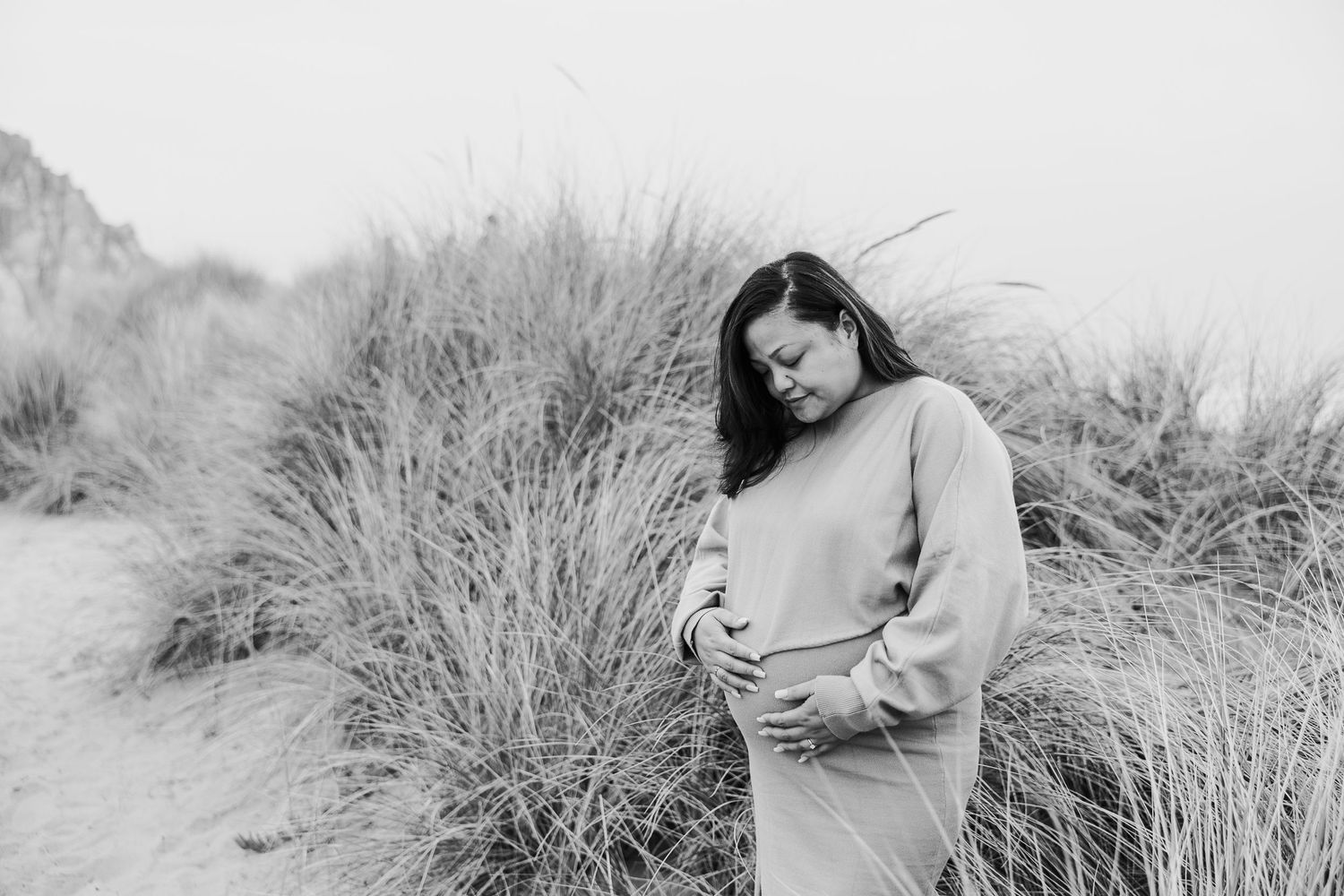 Standing among beach grass and dunes, a person in a cozy sweater looks down thoughtfully.