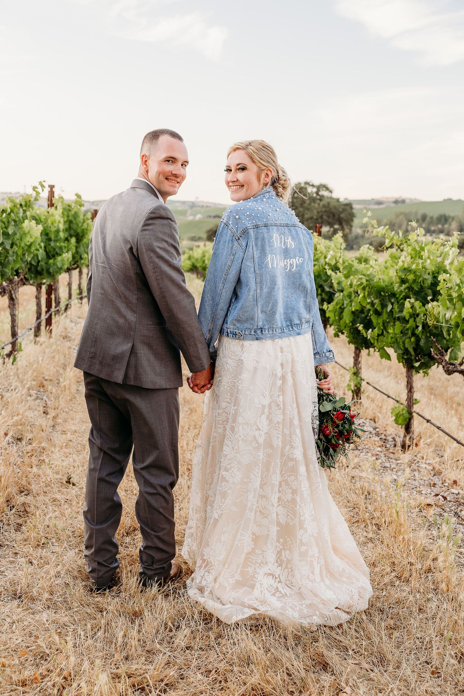 Newlyweds hold hands while showing off a custom denim jacket in a vineyard.