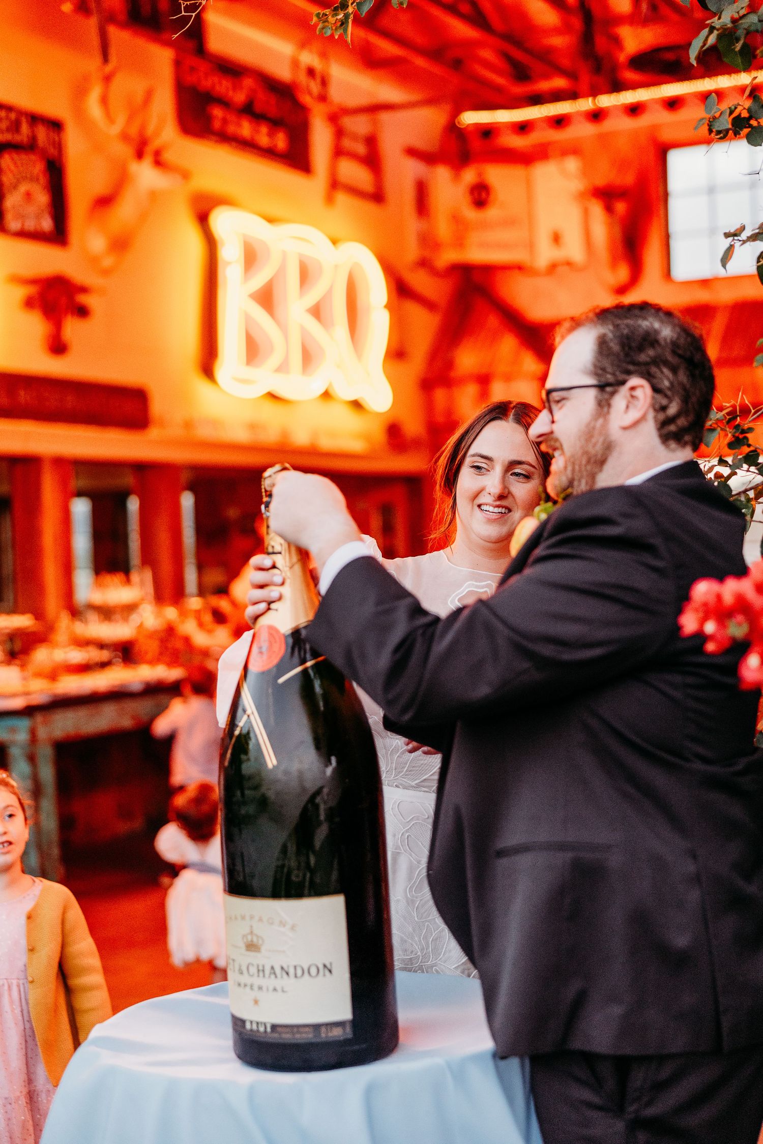 A couple celebrates by opening a large champagne bottle under an orange neon BBQ sign in San Luis Obispo Holland Ranch.