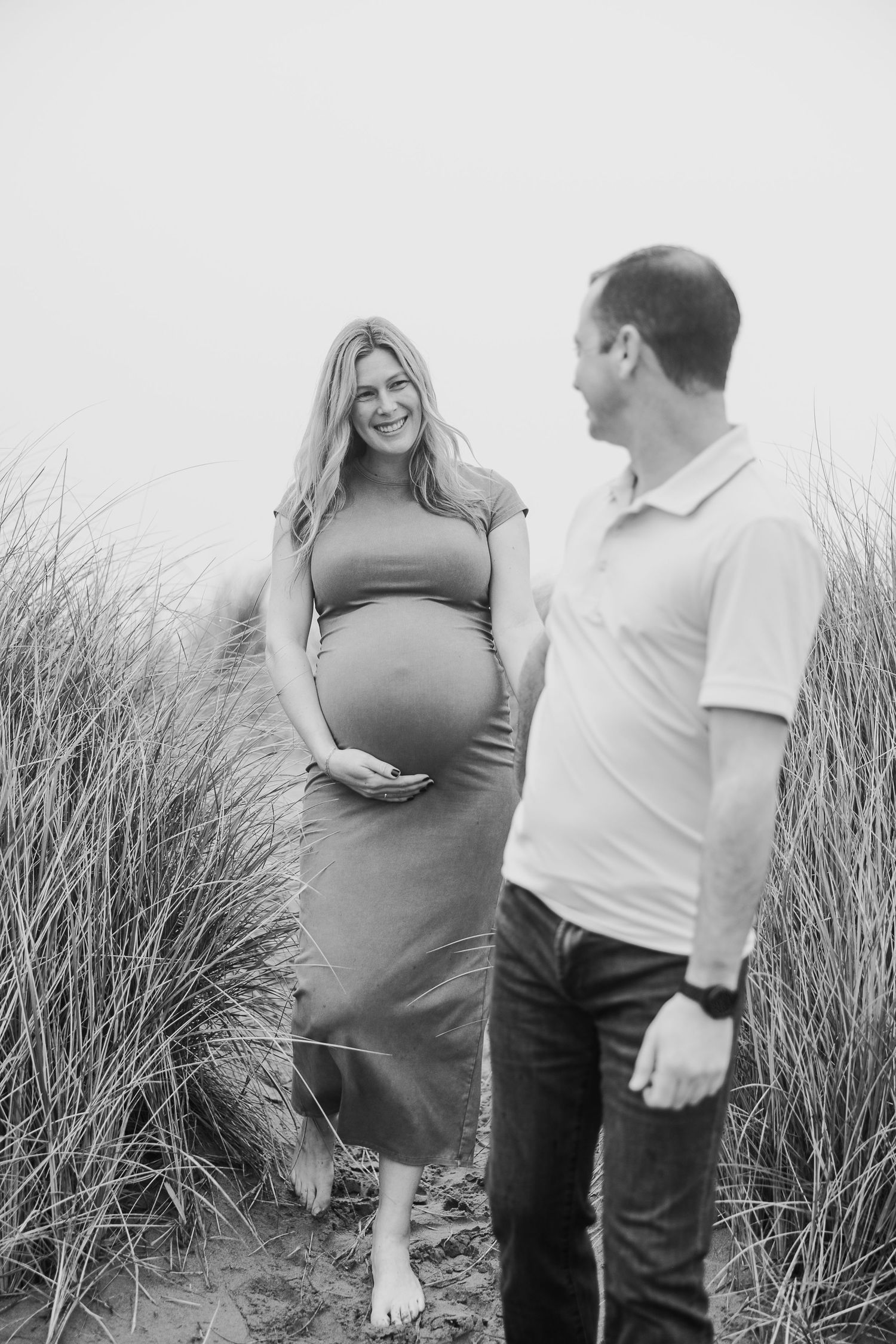 Black and white portrait of an expectant couple sharing a moment among coastal grasses.