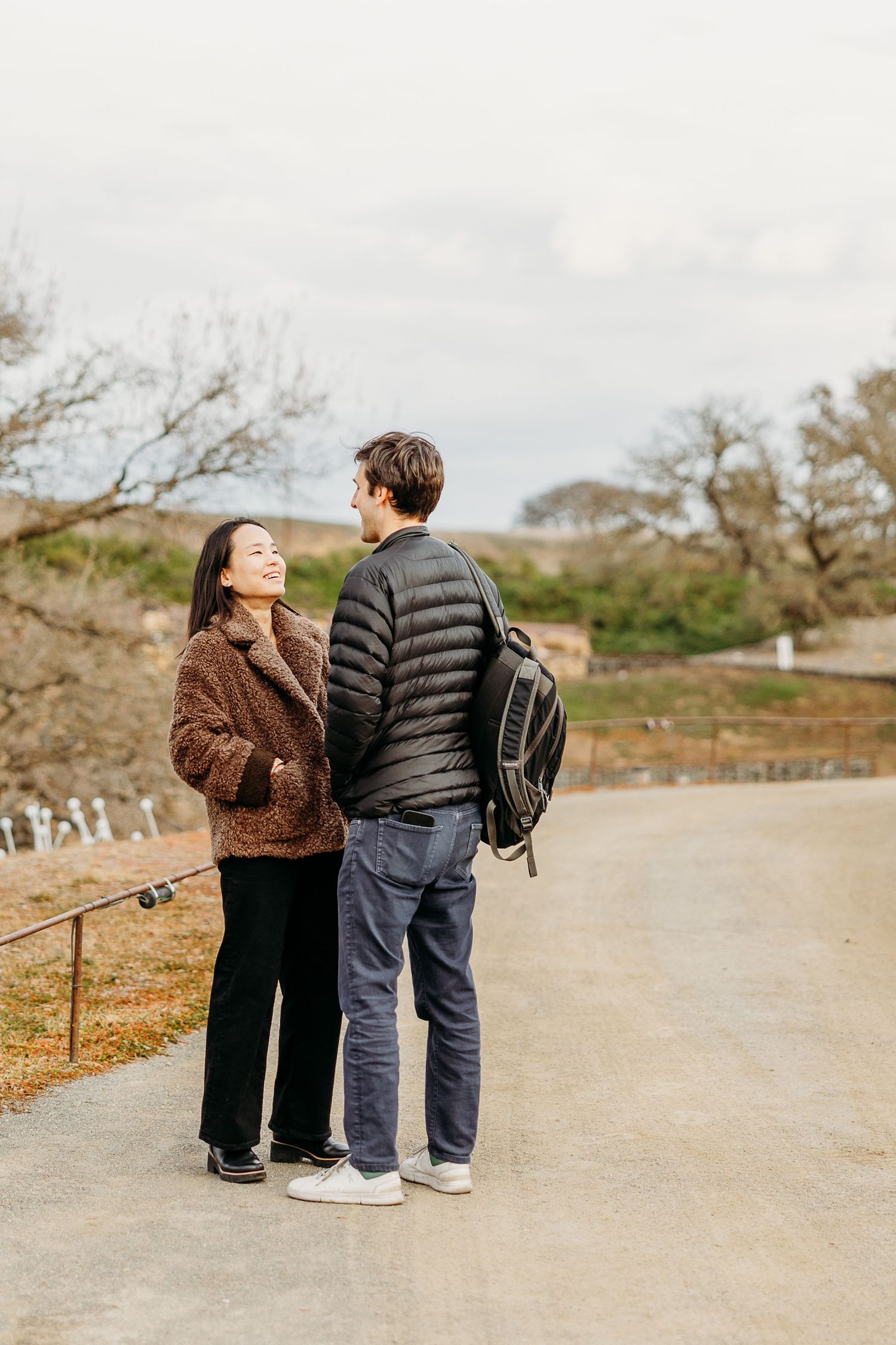 A couple stands together on a path outdoors, wearing casual winter clothing and a backpack on a cloudy day.