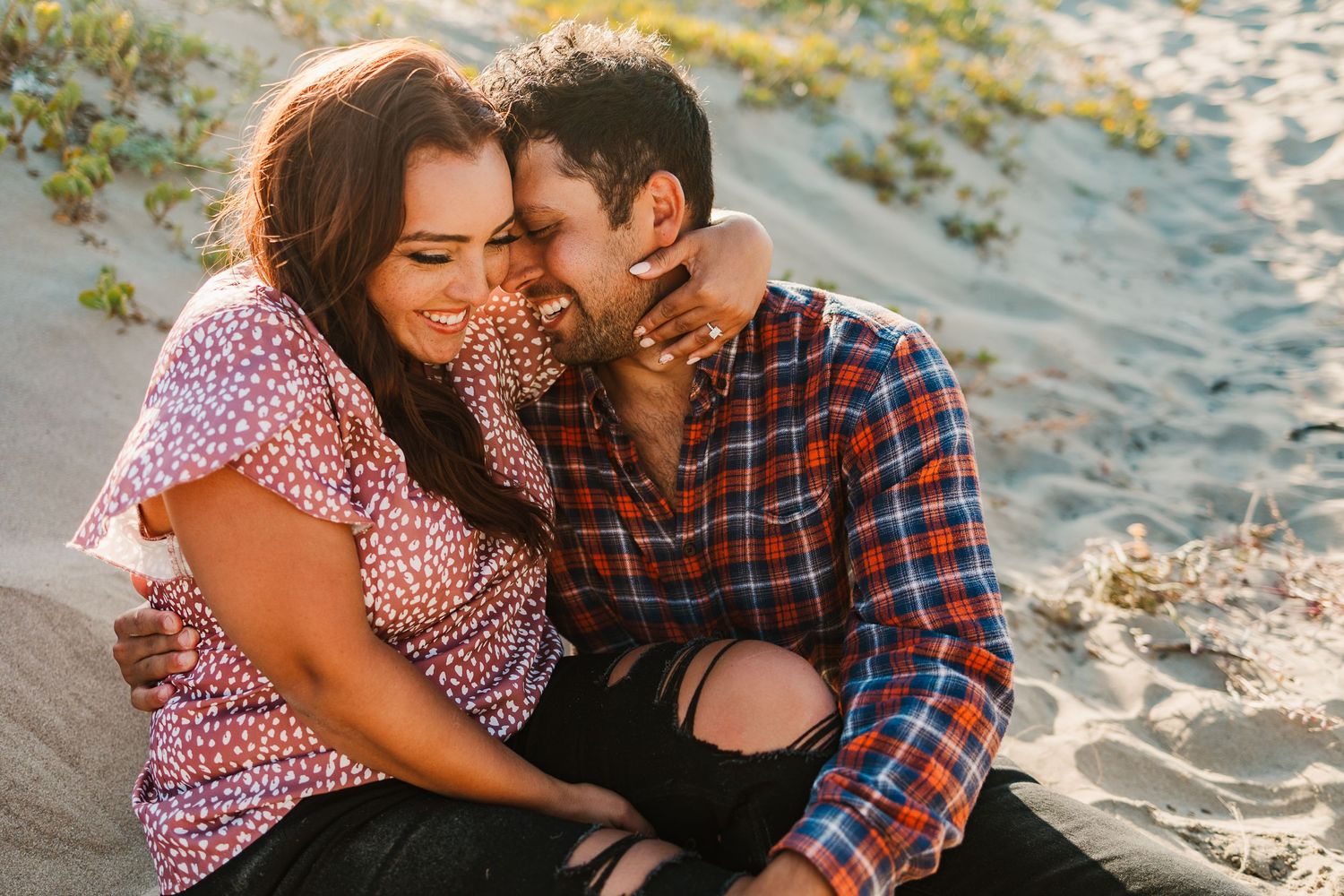Affectionate couple laughing together while sitting on sand dunes in warm evening light.