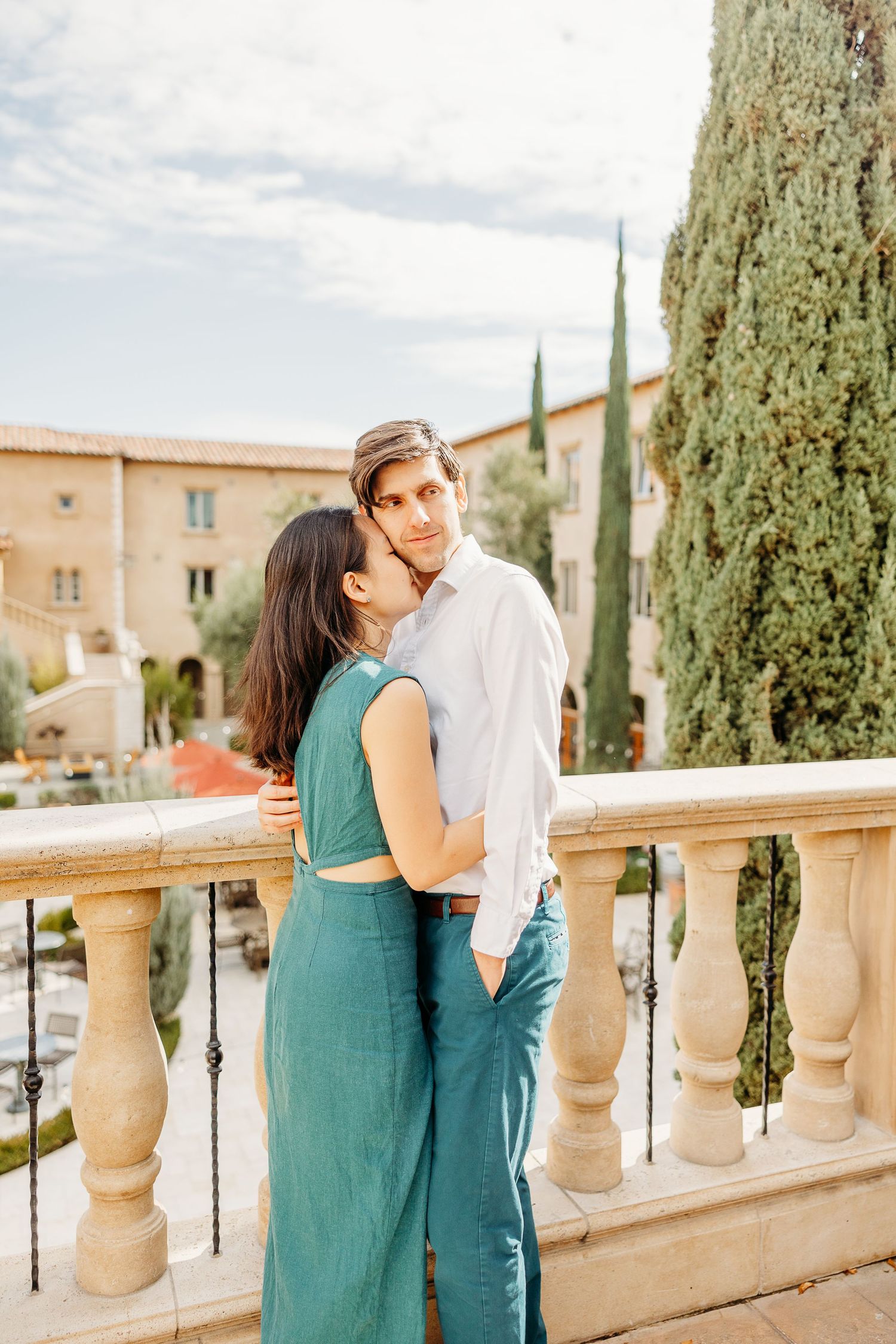 A couple embraces on an elegant balcony with Mediterranean cypress trees in the background.