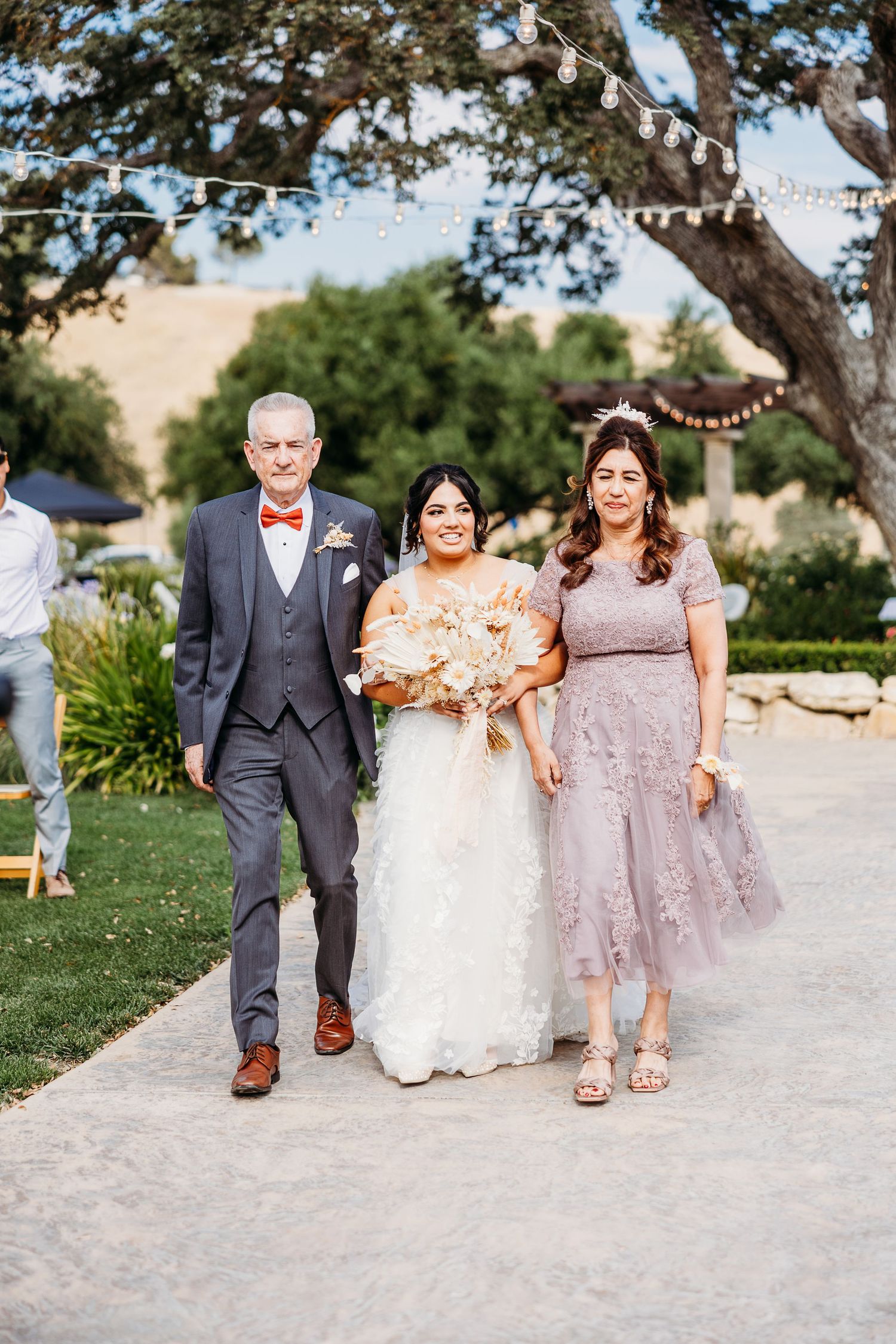 A wedding processional walks along a garden path under string lights and trees at an outdoor venue.