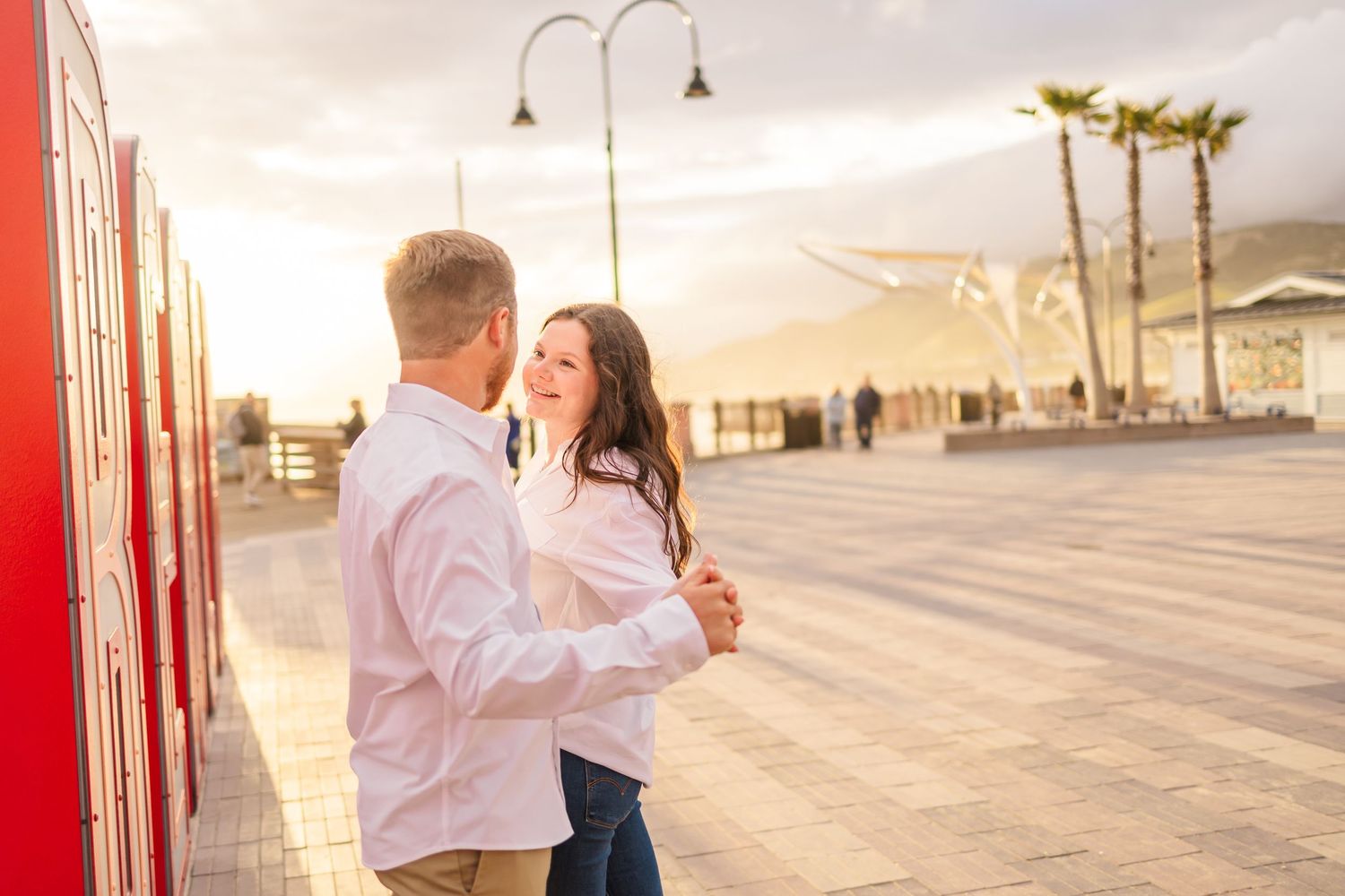 Couple embraces while dancing near red wall during sunset photoshoot.