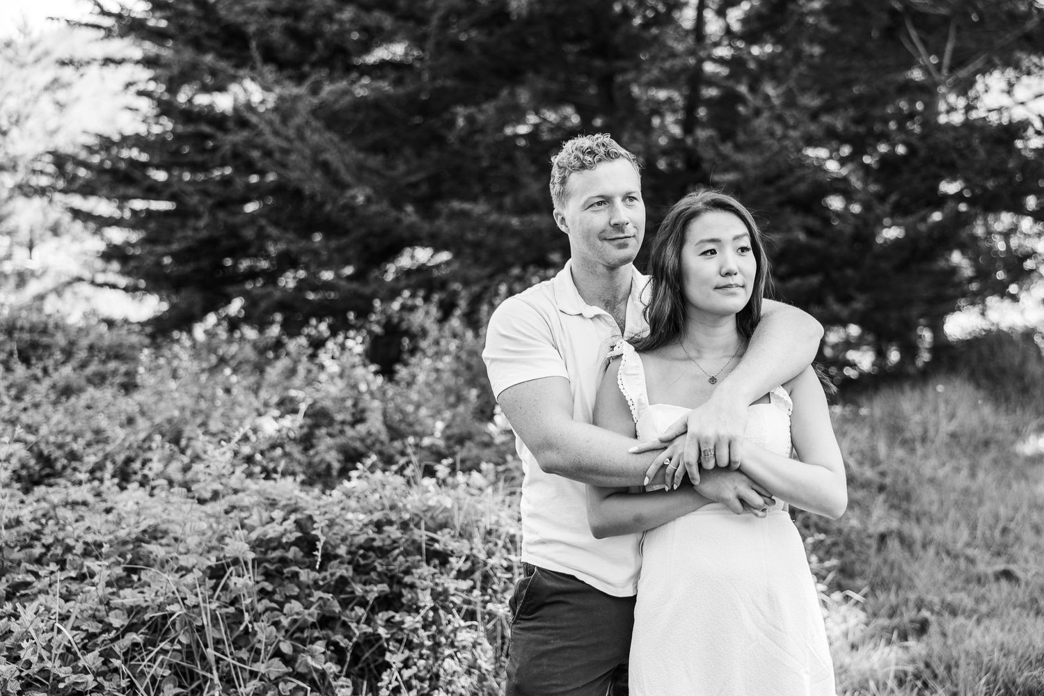 A black and white portrait of a couple embracing outdoors with natural foliage in the background.