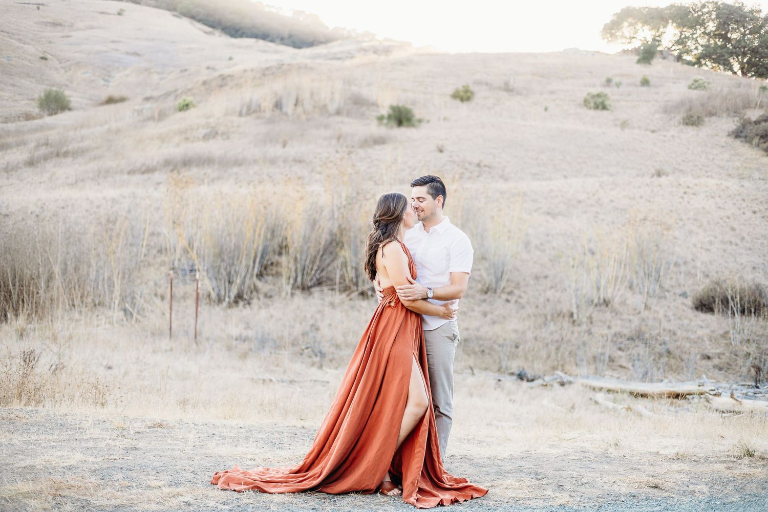 A couple shares an intimate moment on a path with flowing rust-colored fabric catching the breeze.