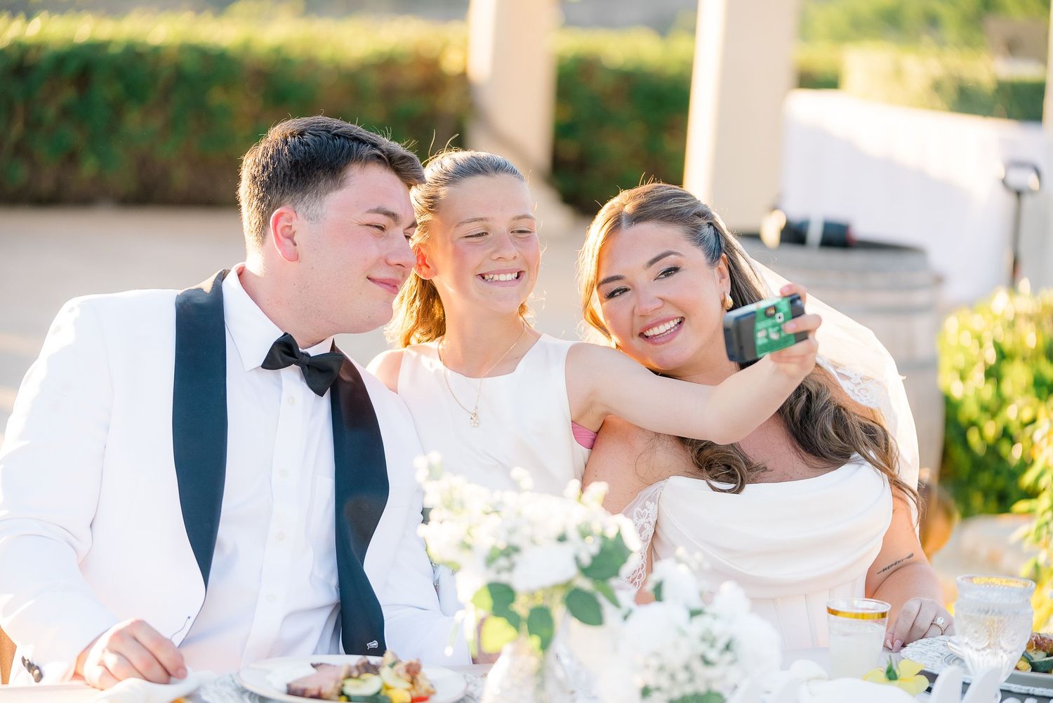 Bride and groom take a selfie with a guest at an outdoor wedding reception table with white floral arrangements.