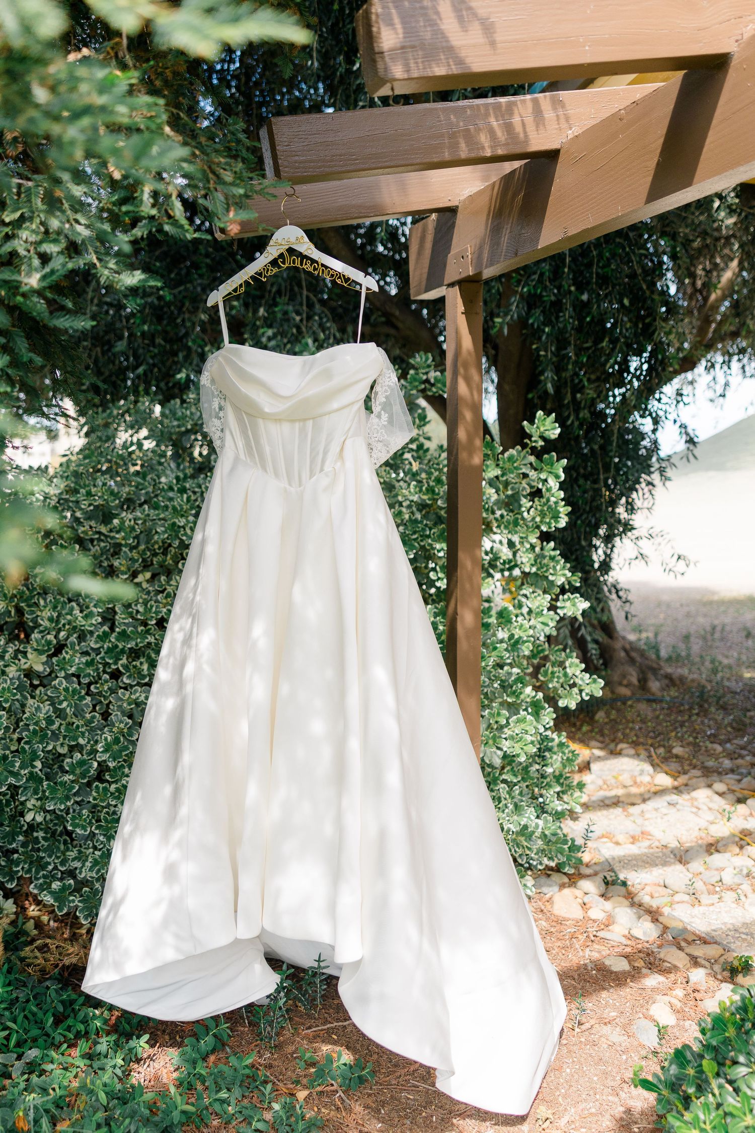 Elegant white off-shoulder wedding gown hanging on a wooden arbor surrounded by lush green trees.